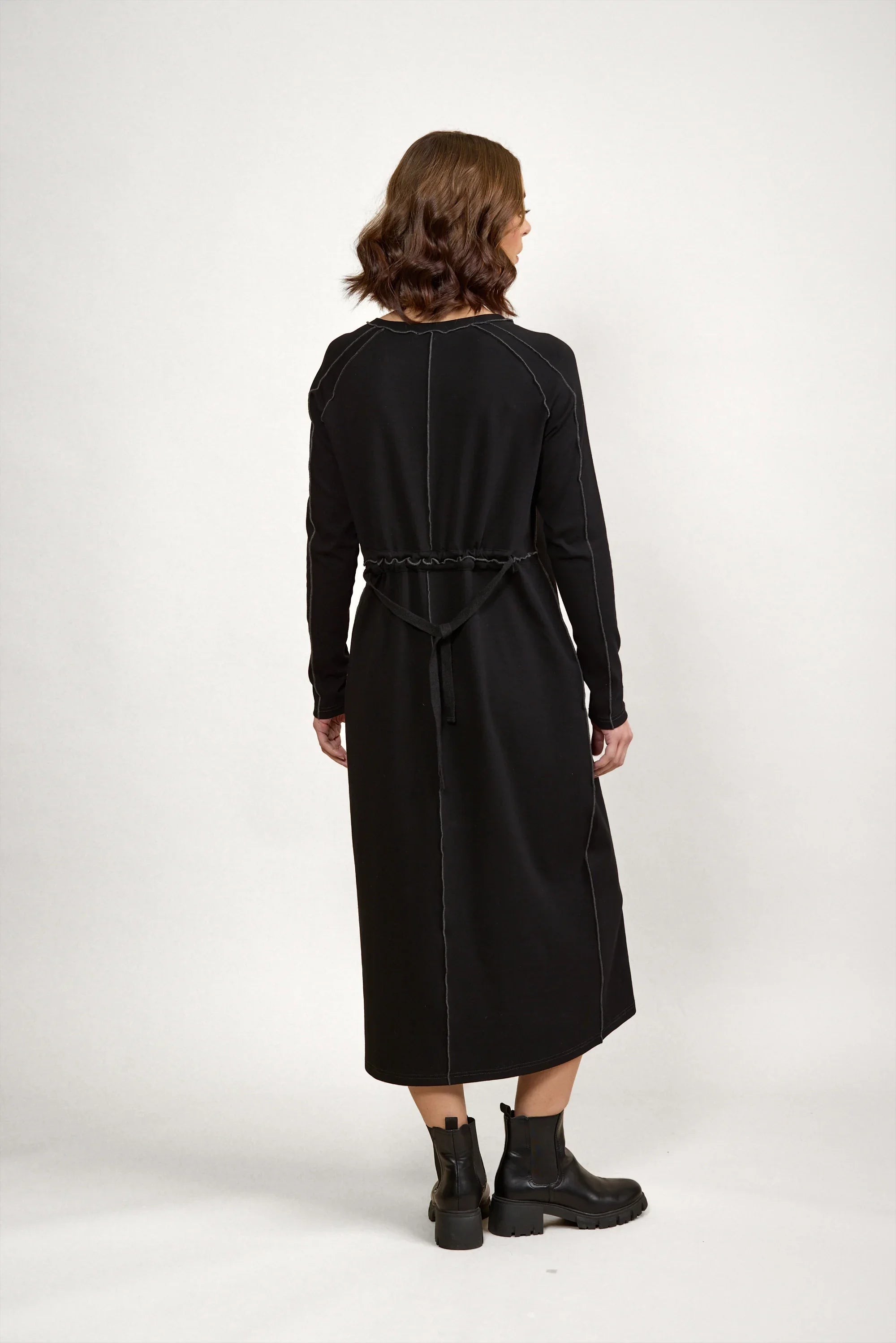 Woman in long sleeve black midi dress with visible seam details, back tie, and ankle boots