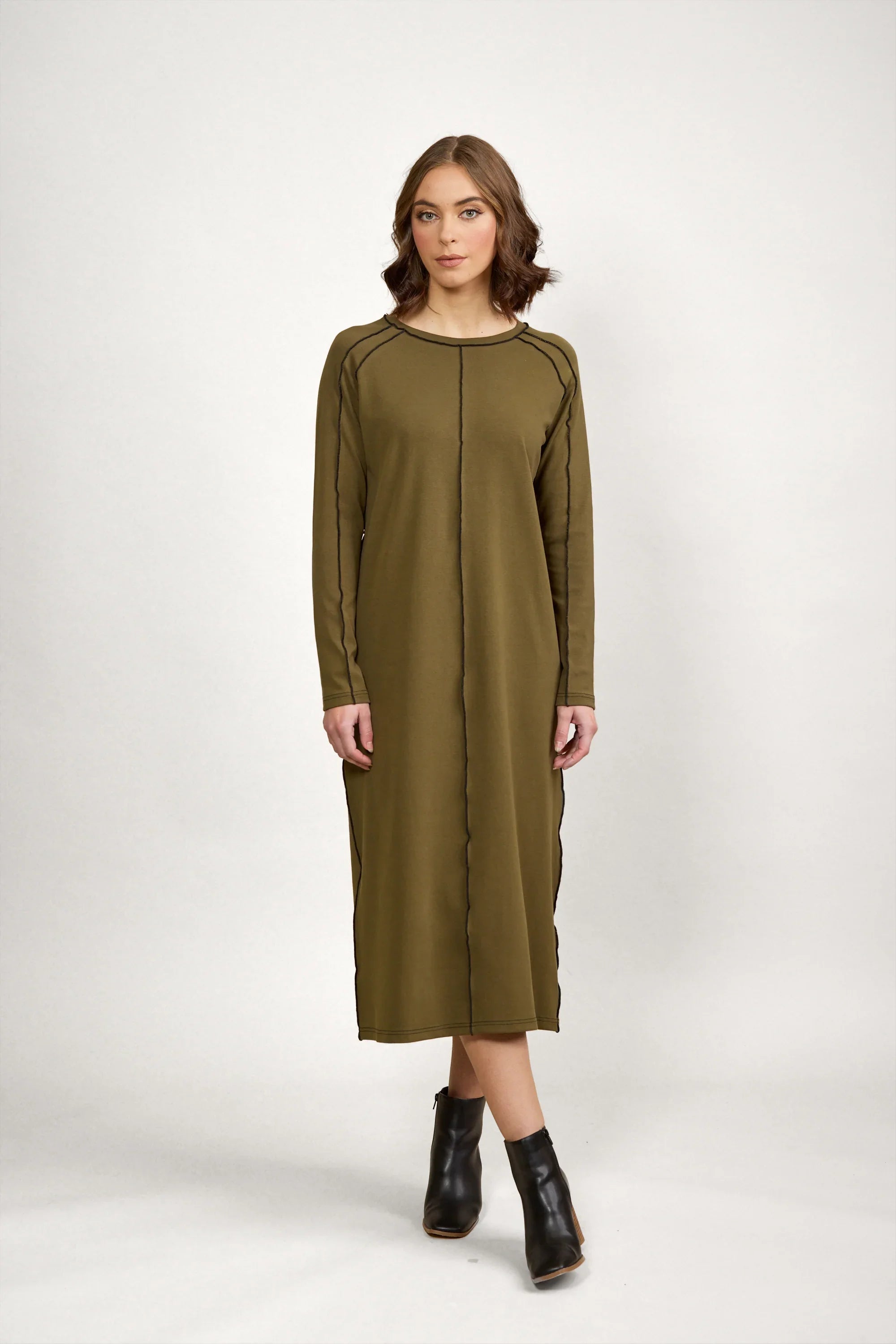 Woman wearing olive green long sleeve midi dress with black seam details and black ankle boots, standing against a plain white background.