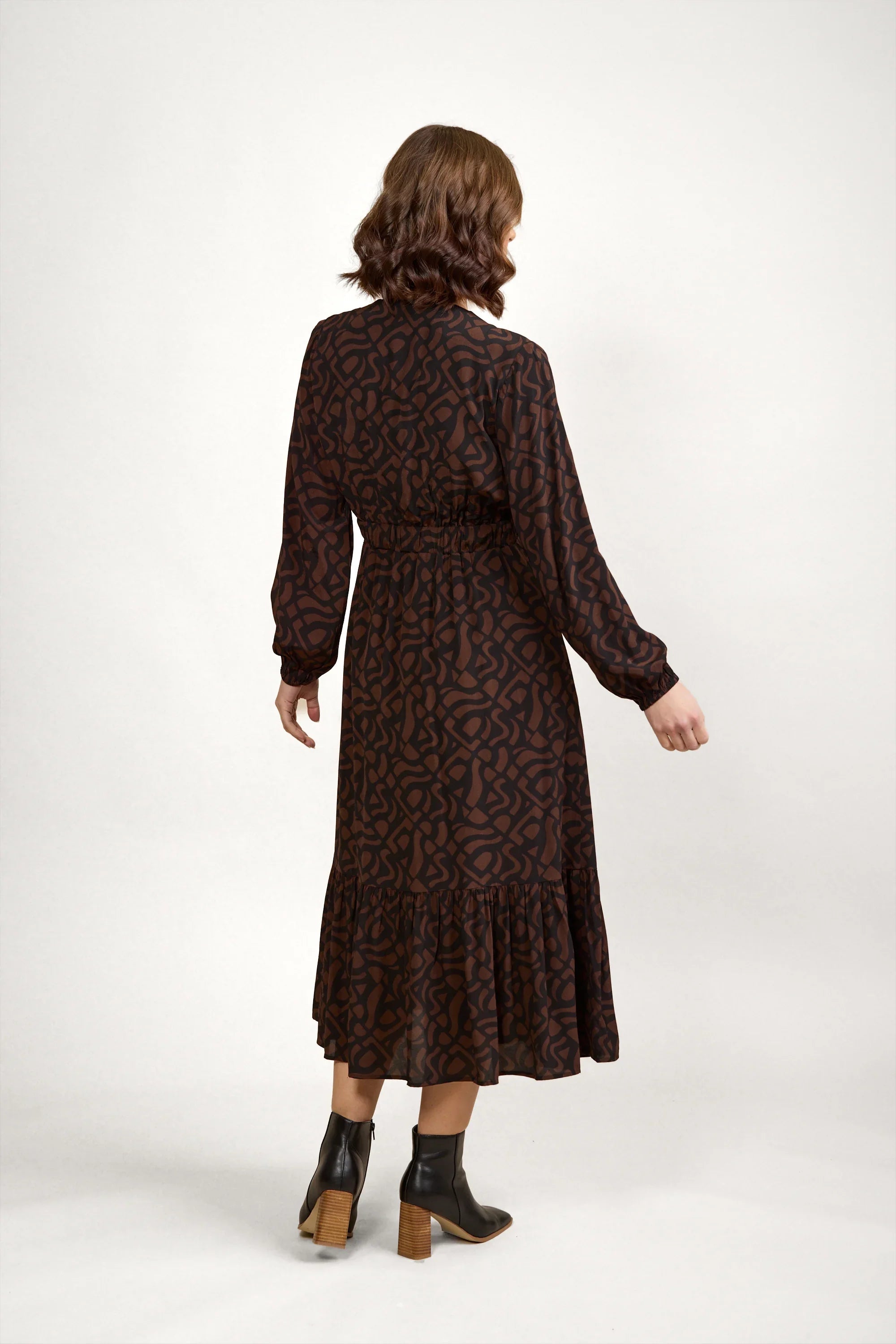 Woman modeling long sleeve mocha printed midi dress with ruffled hem and ankle boots, back view