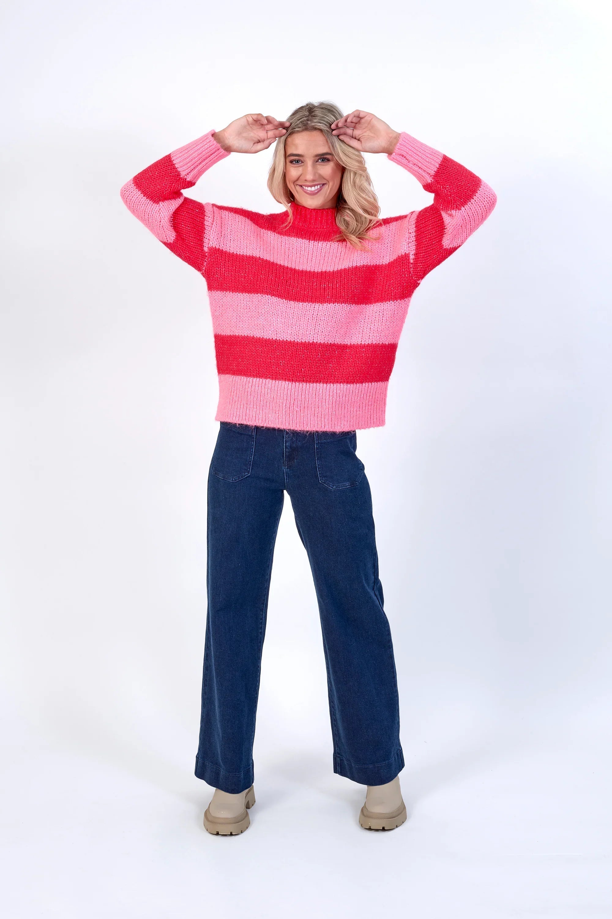 Woman wearing pink and red striped knit sweater with blue wide-leg jeans, studio background
