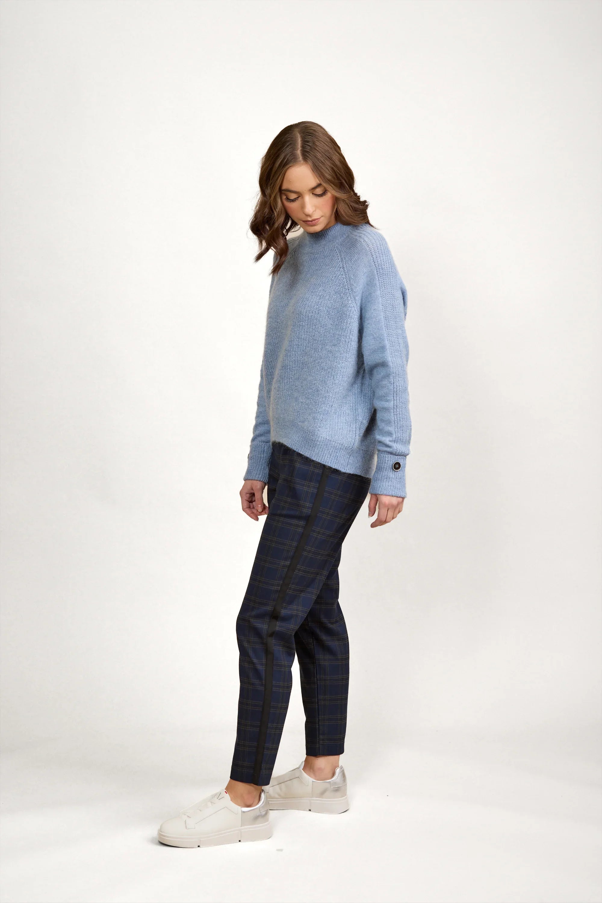Woman wearing a sky blue knit sweater, plaid pants, and white sneakers against a plain background.