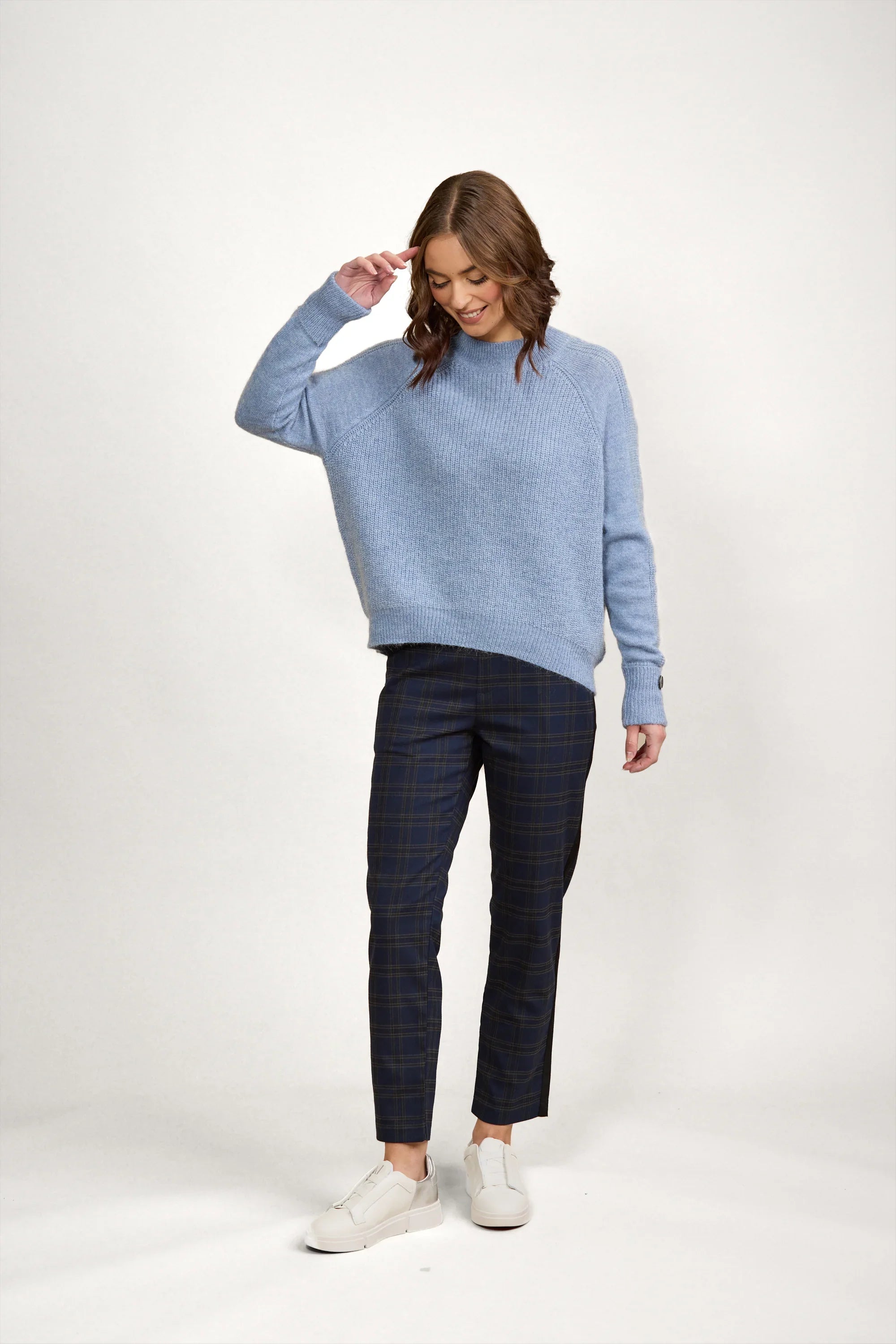 Woman wearing a sky blue knit sweater, plaid pants, and white sneakers in studio setting