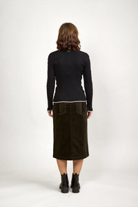 Woman facing away in black ribbed sweater, olive midi skirt, ankle boots, neutral background