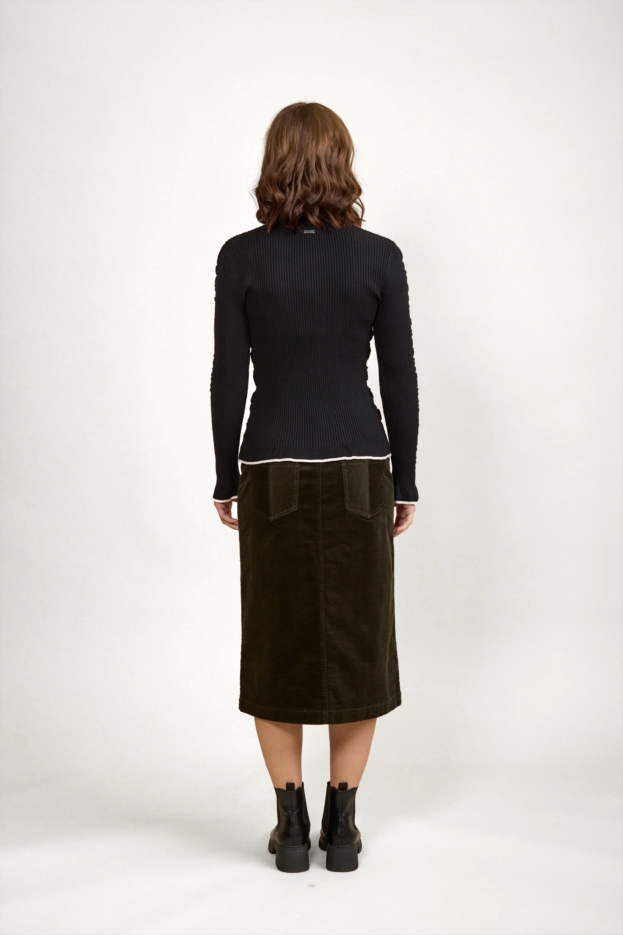 Woman facing away in black ribbed sweater, olive midi skirt, ankle boots, neutral background