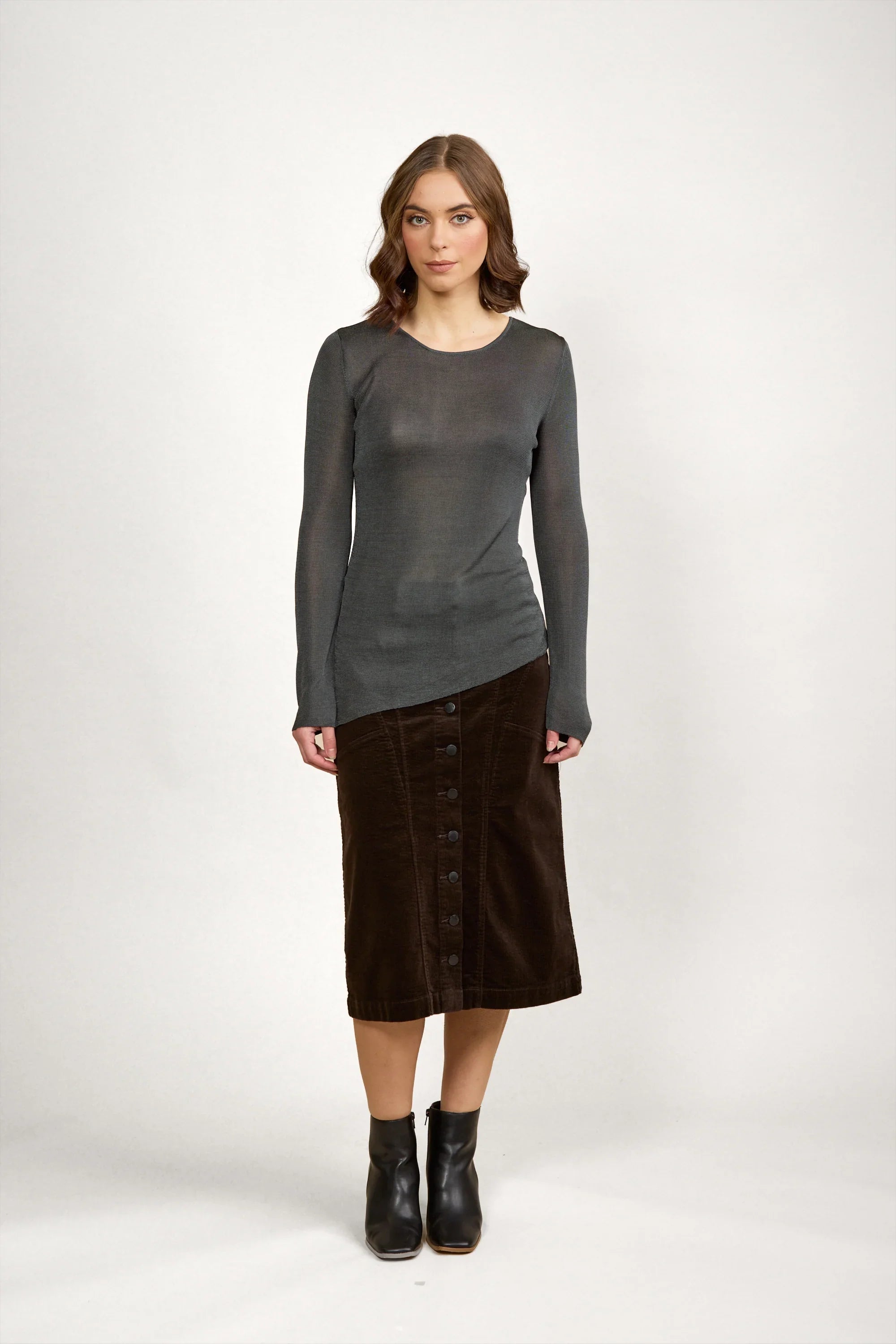 Woman modeling a sheer grey knit sweater with long sleeves, brown button-front midi skirt, and black ankle boots, standing against a white background.