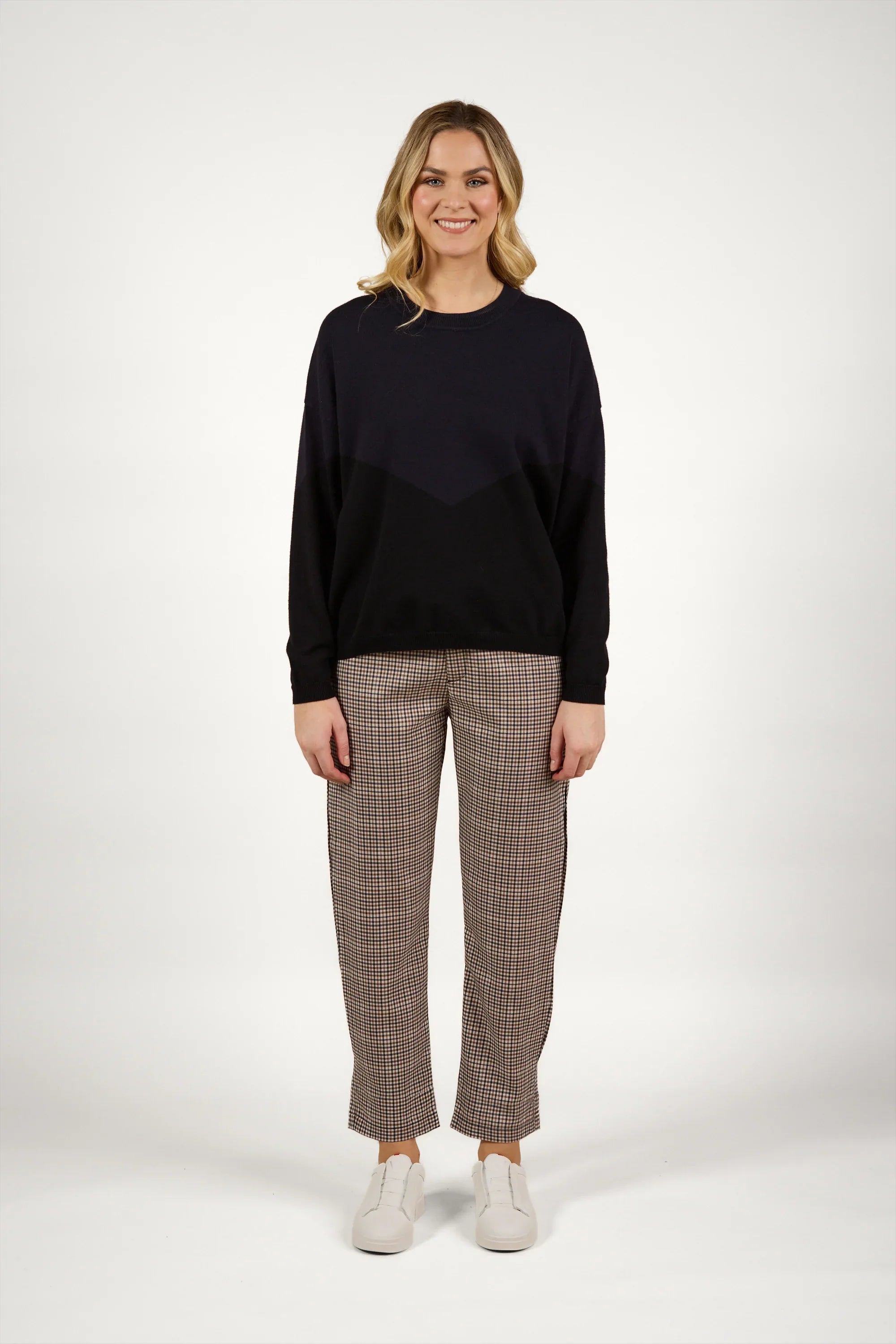 Woman wearing a black knit sweater with checkered pants and white sneakers standing against a plain background.