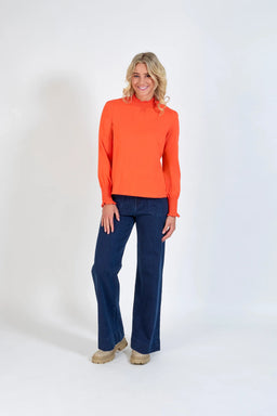 Woman modeling bright orange long sleeve top with ruffled neck and cuffs, wide leg jeans