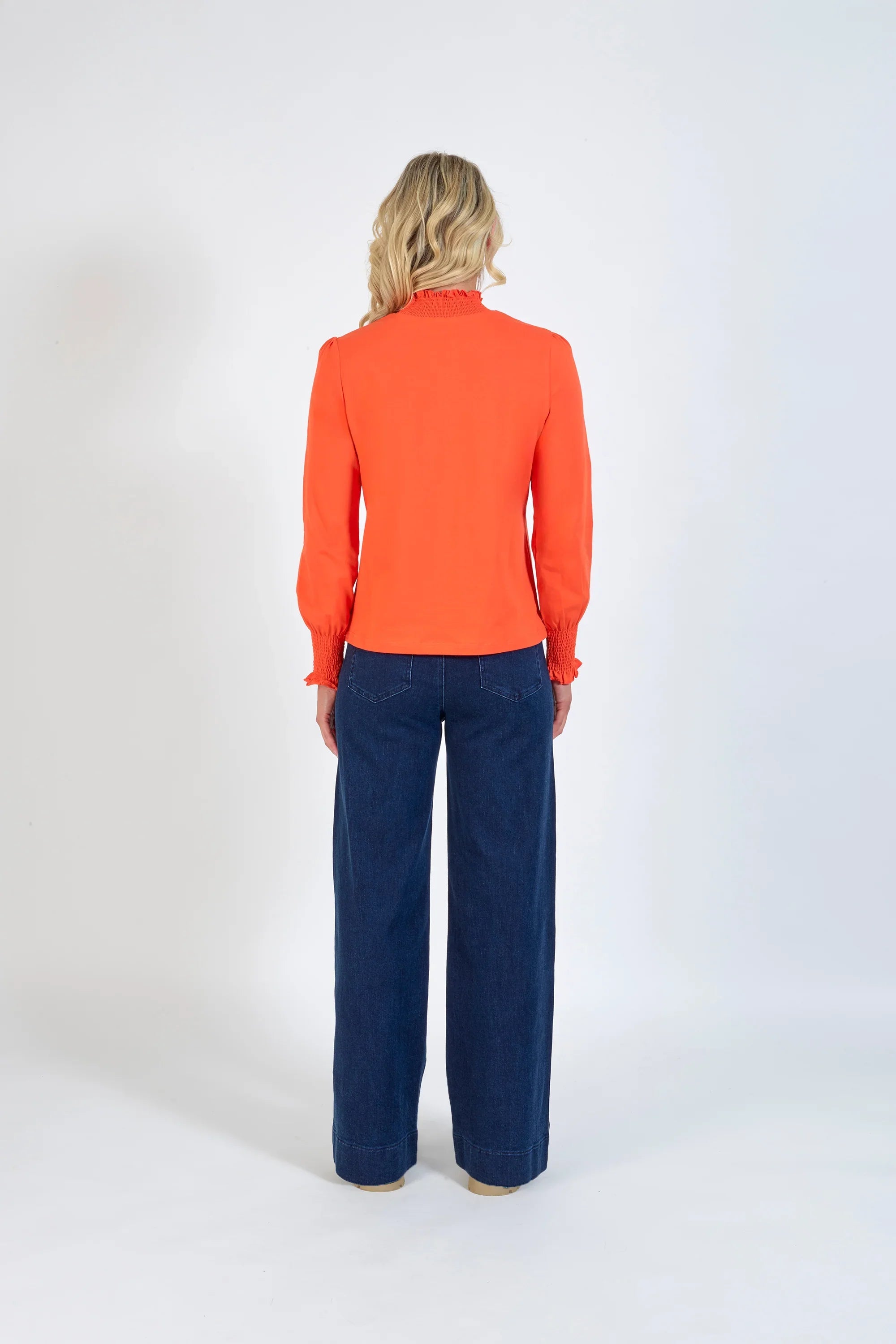 Woman in bright orange long sleeve top with smocked cuffs and dark blue wide-leg jeans, back view