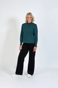 Woman wearing a dark green long sleeve top with smocked cuffs and neck, black wide leg pants, and white sneakers, standing against a plain background.