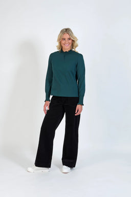 Woman wearing a dark green long sleeve top with smocked cuffs and neck, black wide leg pants, and white sneakers, standing against a plain background.