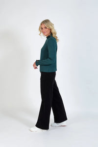 Woman in teal long sleeve top with shirred cuffs and collar, black pants, and white sneakers