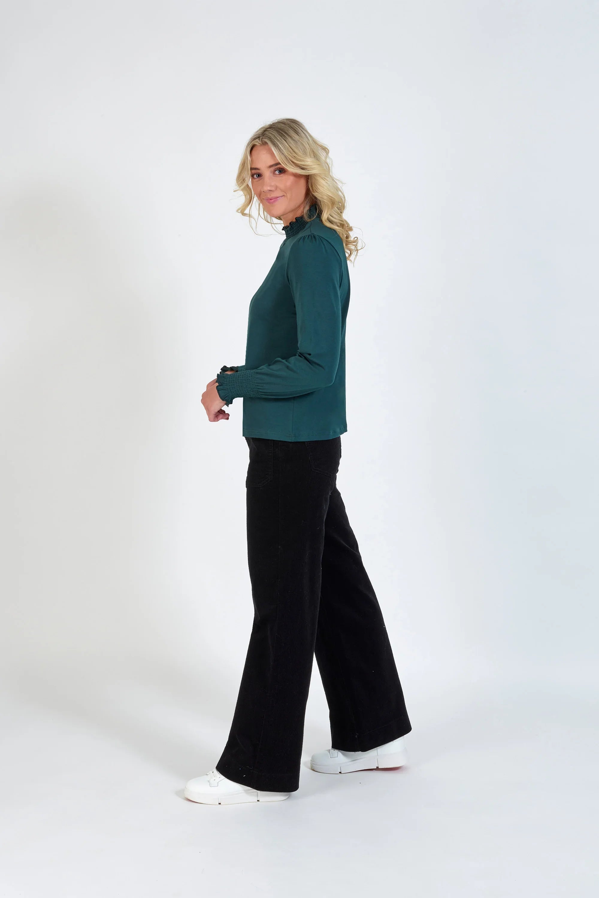 Woman in teal long sleeve top with shirred cuffs and collar, black pants, and white sneakers