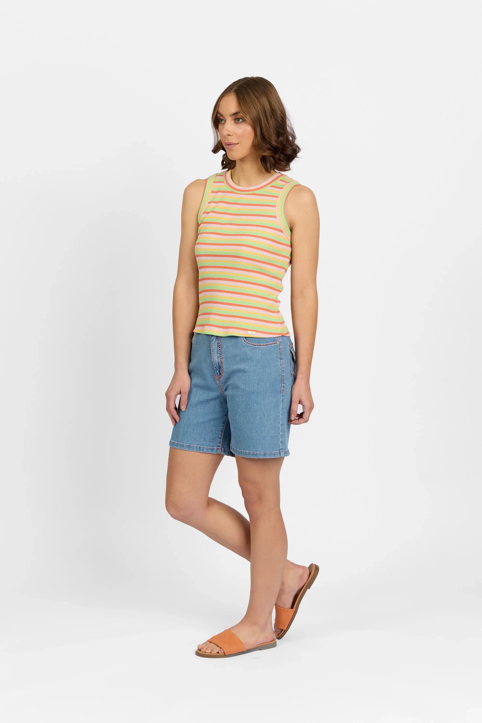 Woman wearing a colorful striped tank top, denim shorts, and orange sandals against white background