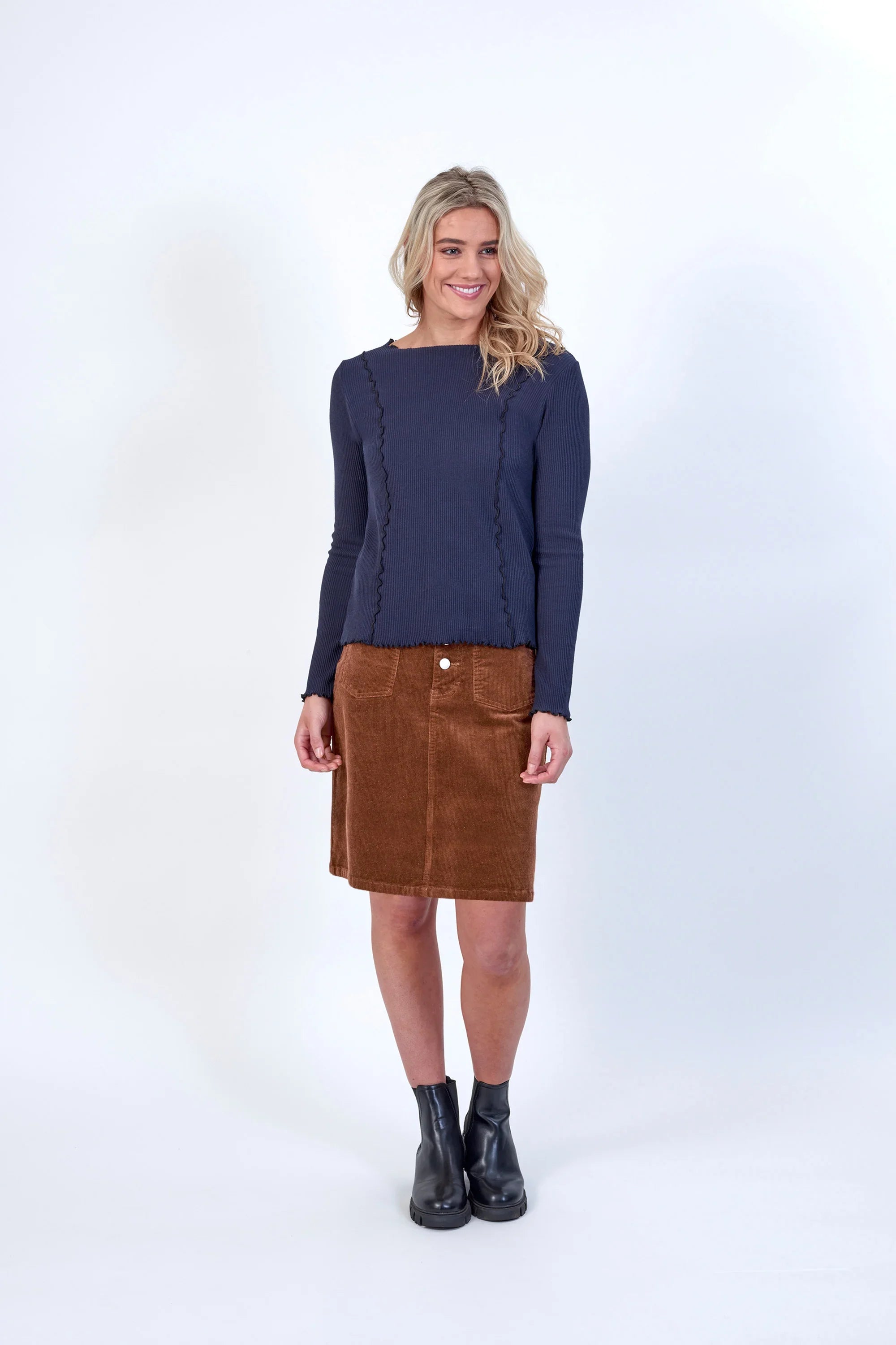 Woman wearing navy ribbed long sleeve top with wavy trim, brown corduroy skirt, black boots