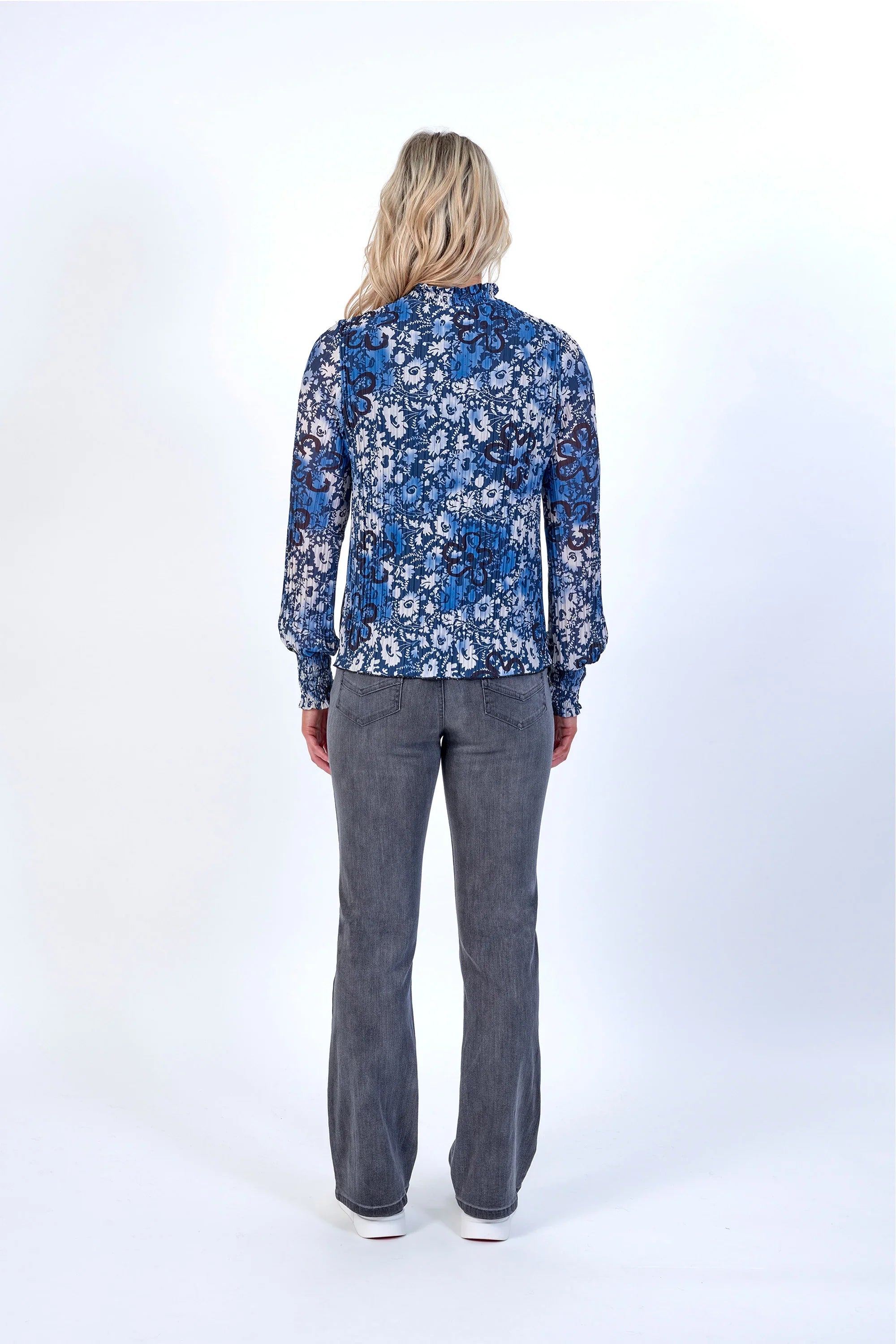Woman in long sleeve blue floral blouse and grey jeans, standing with back to camera