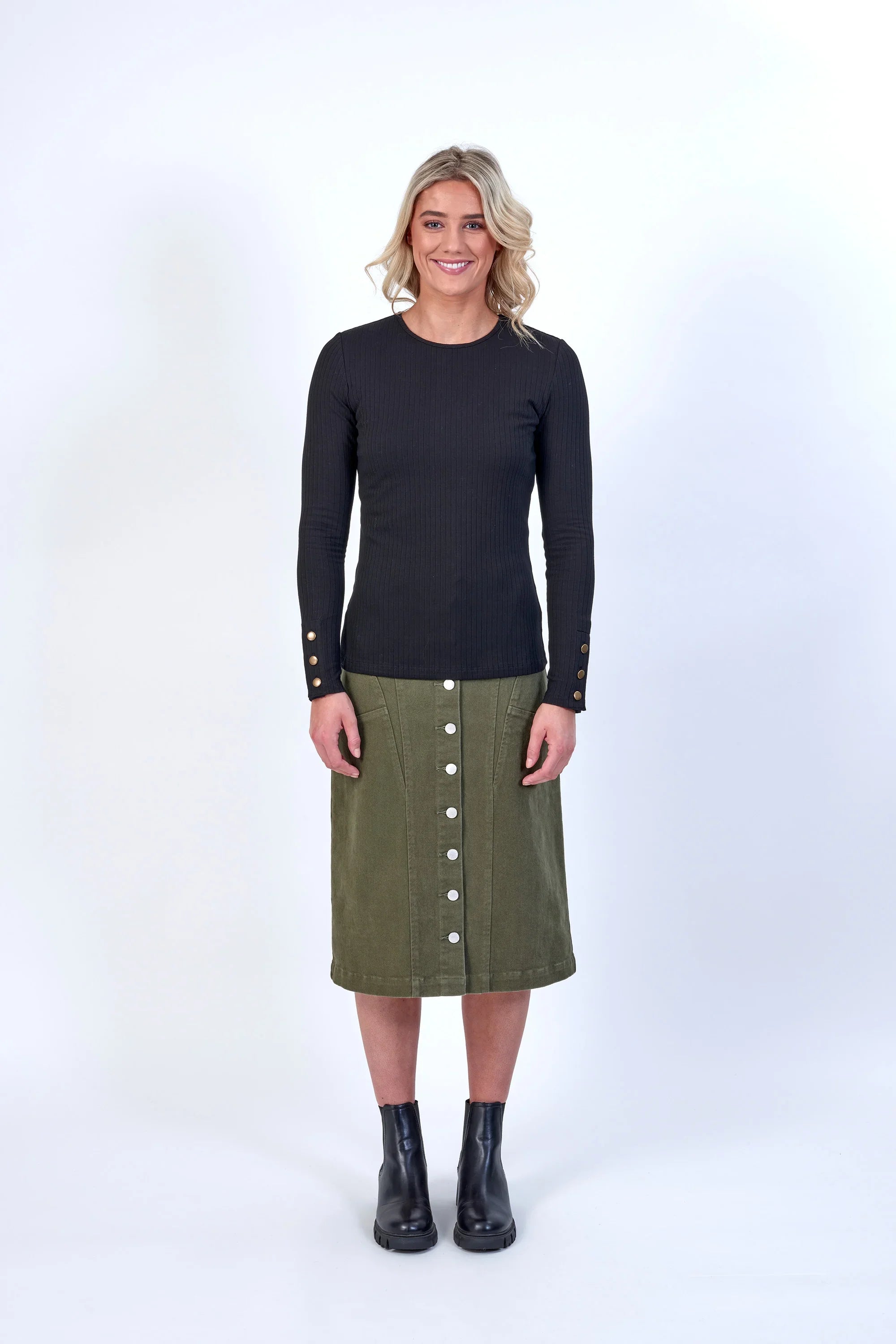 Woman wearing a black ribbed long sleeve top with gold button cuffs, olive green midi skirt with front buttons, and black ankle boots, standing in a studio with a white background.
