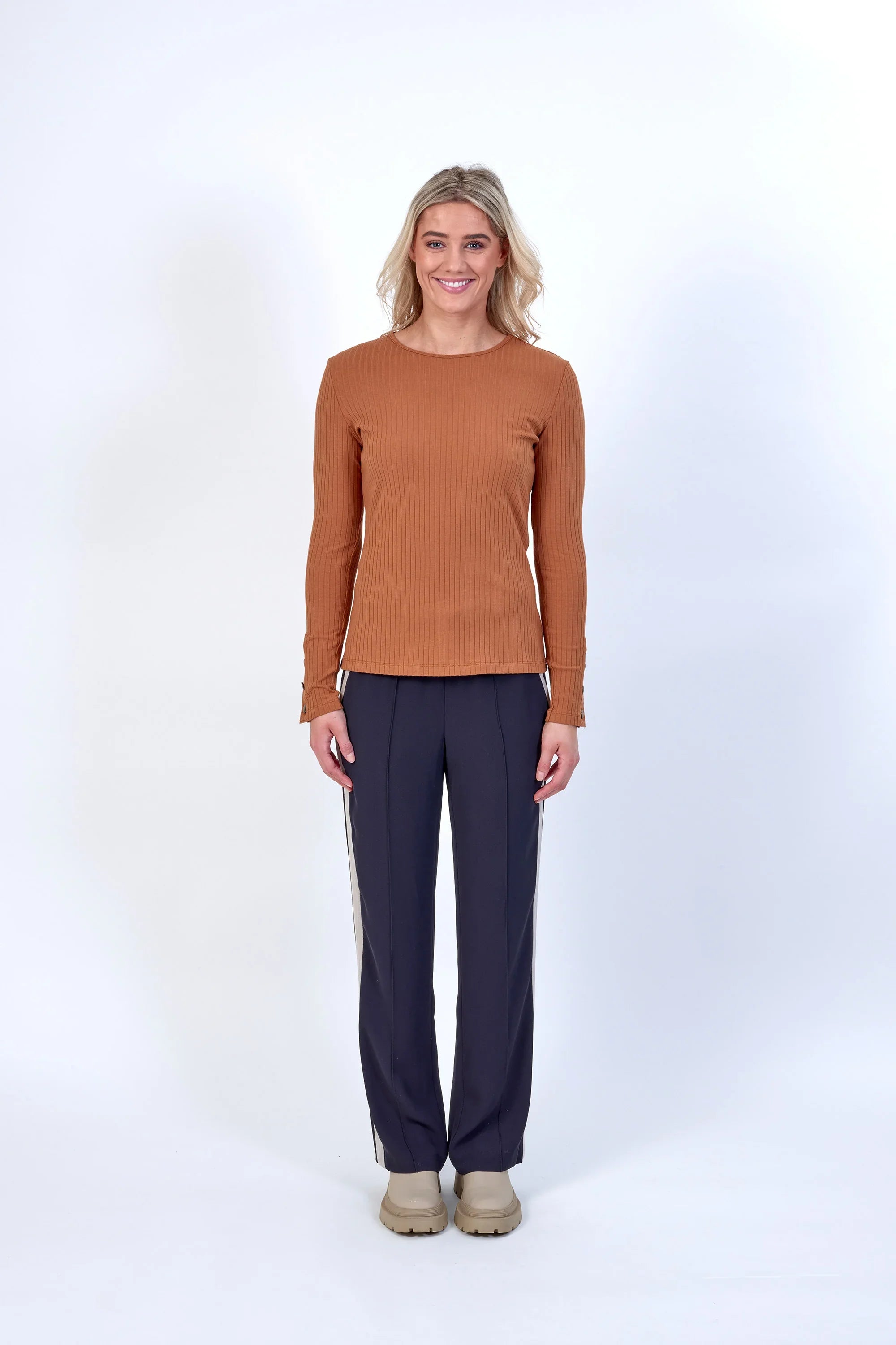 Woman wearing a ribbed long sleeve brown top with dark pants, standing against white background