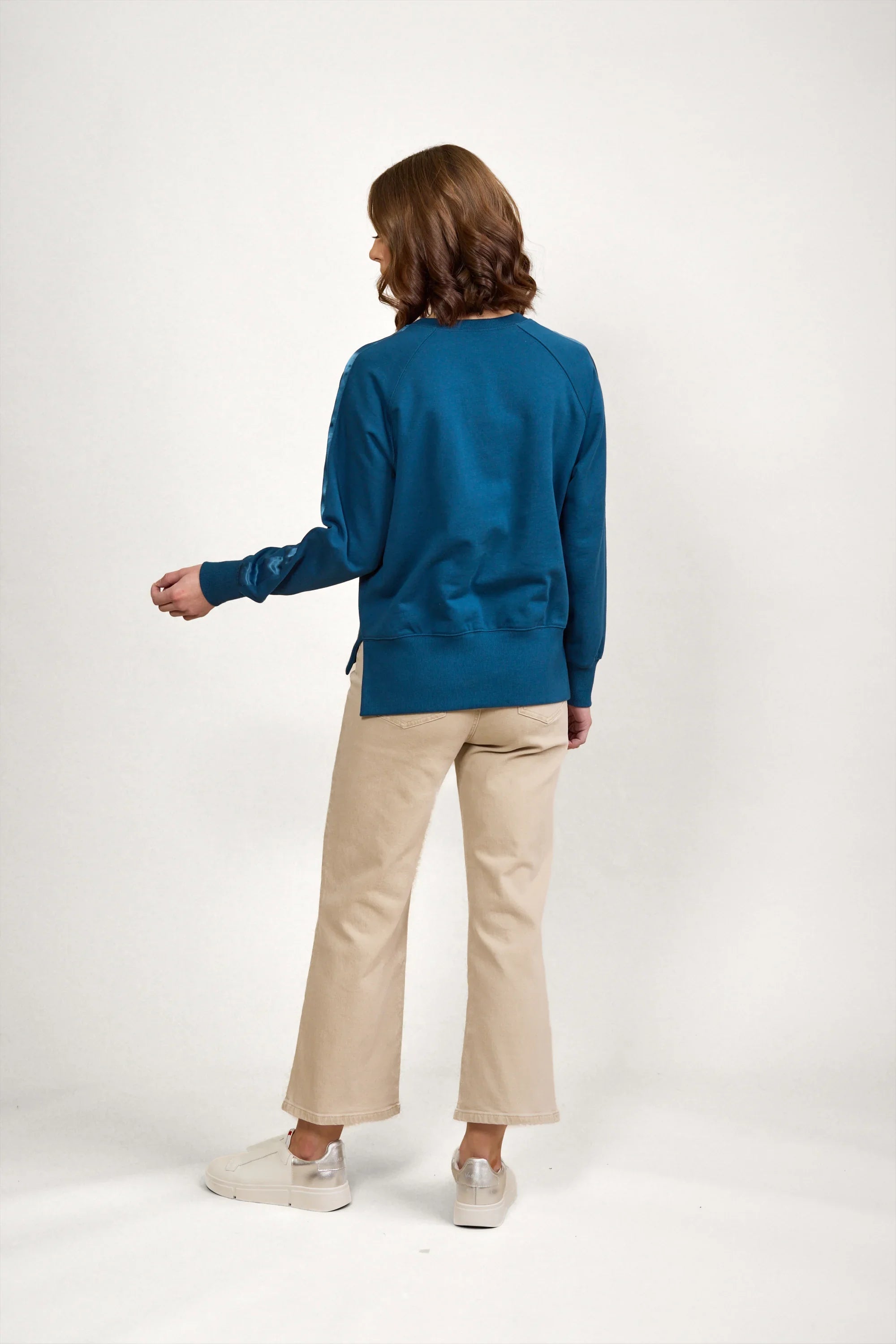 Woman wearing teal knit jumper with beige trousers and white sneakers, back view