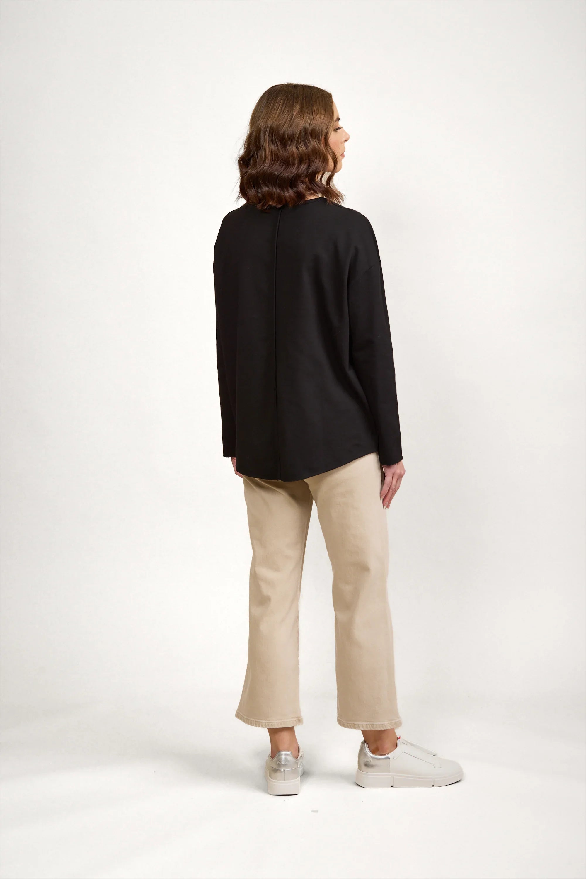 Woman wearing black long sleeve top and beige pants, viewed from the back, in studio