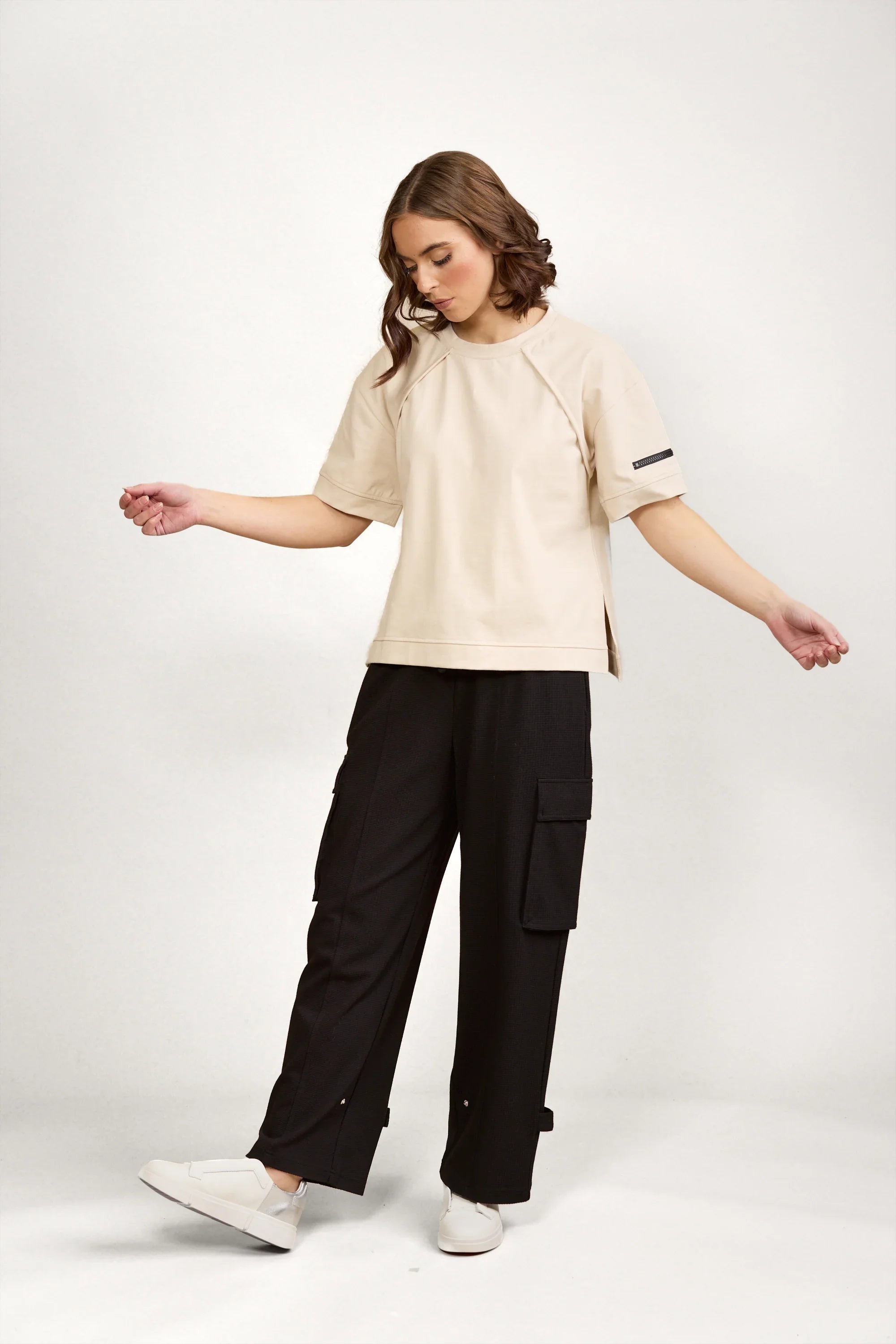 Woman in a beige tee and black cargo pants with white sneakers, minimalist studio backdrop.