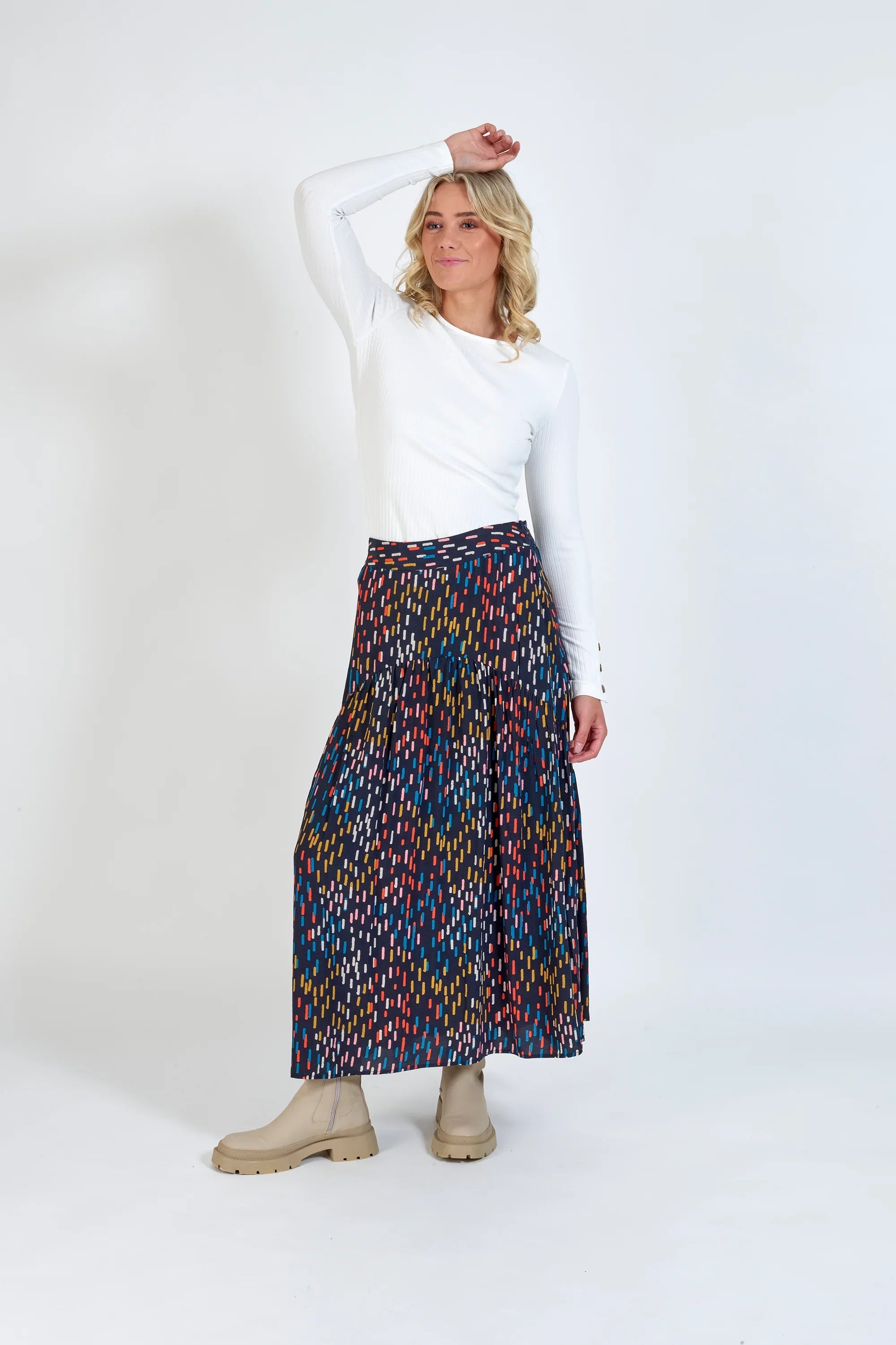 Woman in white long sleeve top, colorful patterned pleated maxi skirt, and beige boots