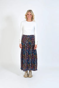 Woman in a white long-sleeve top and colorful patterned maxi skirt, standing indoors