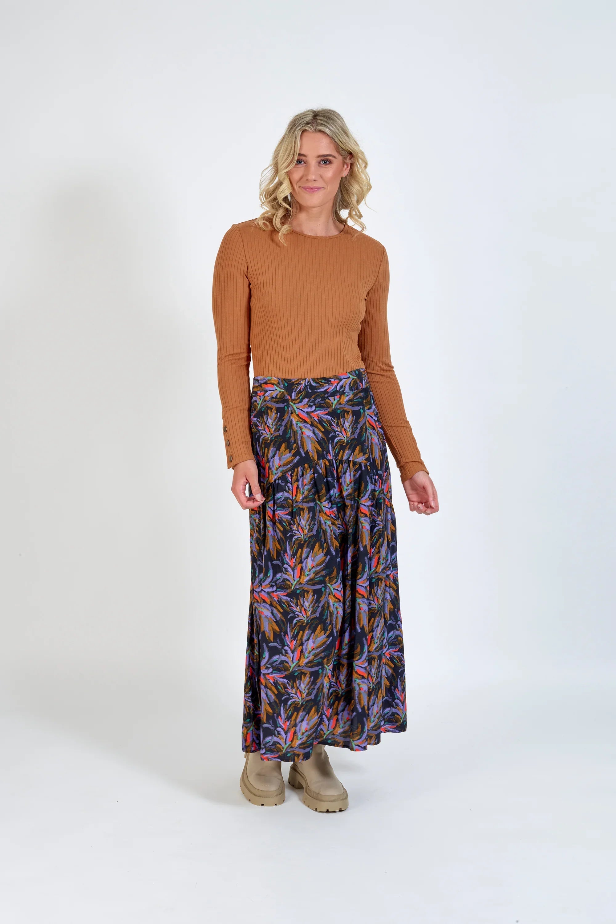 Woman wearing a tan ribbed knit top and colorful floral maxi skirt, neutral background