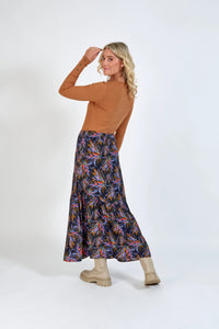 Woman wearing a long, colorful floral skirt and tan ribbed top with light boots, studio background