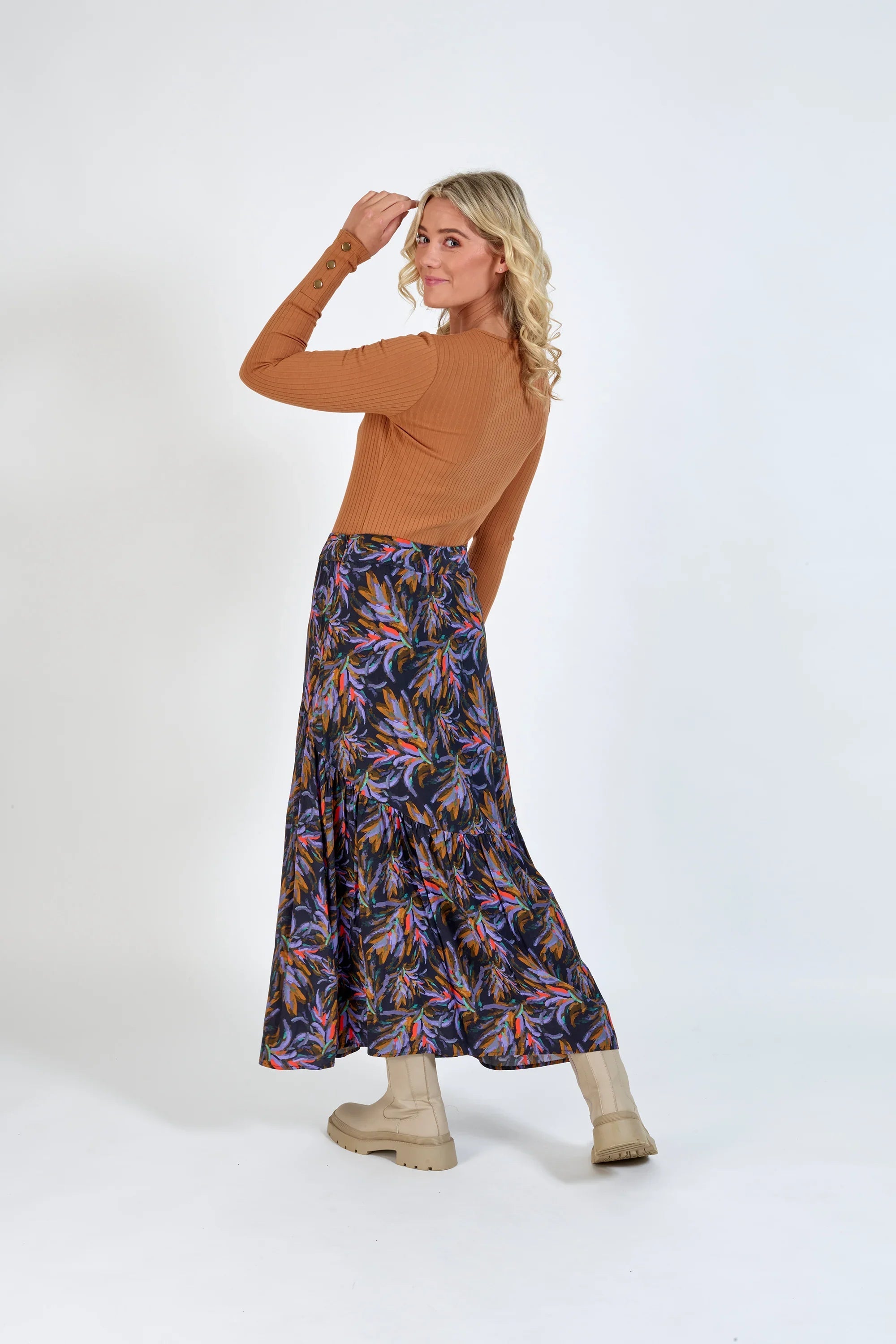 Woman wearing a long, colorful floral skirt and tan ribbed top with light boots, studio background