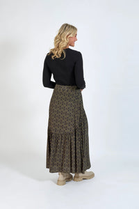 Woman wearing a long geometric print skirt, black ribbed top, and beige boots, standing against a plain background.