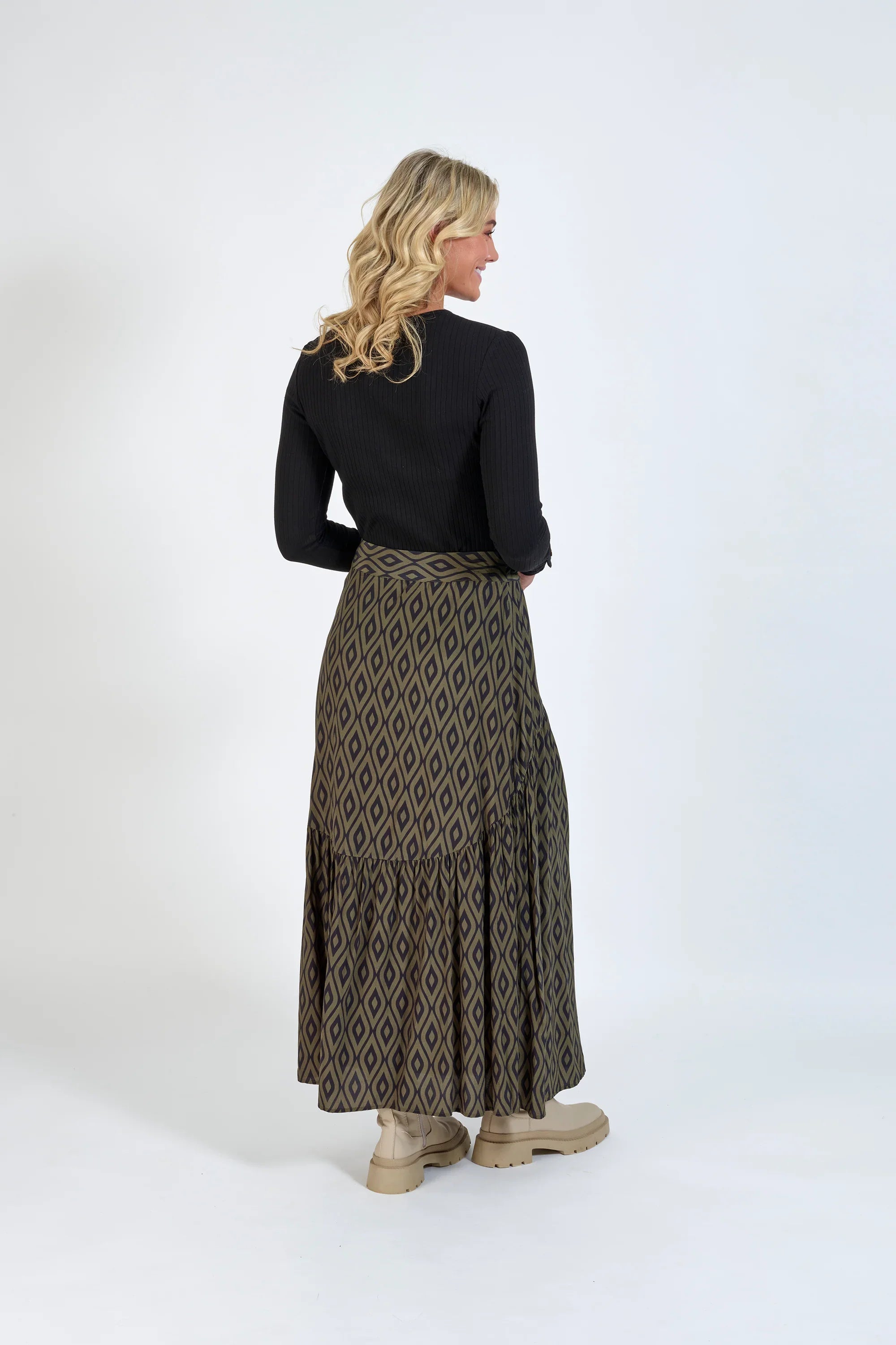 Woman wearing a long geometric print skirt, black ribbed top, and beige boots, standing against a plain background.