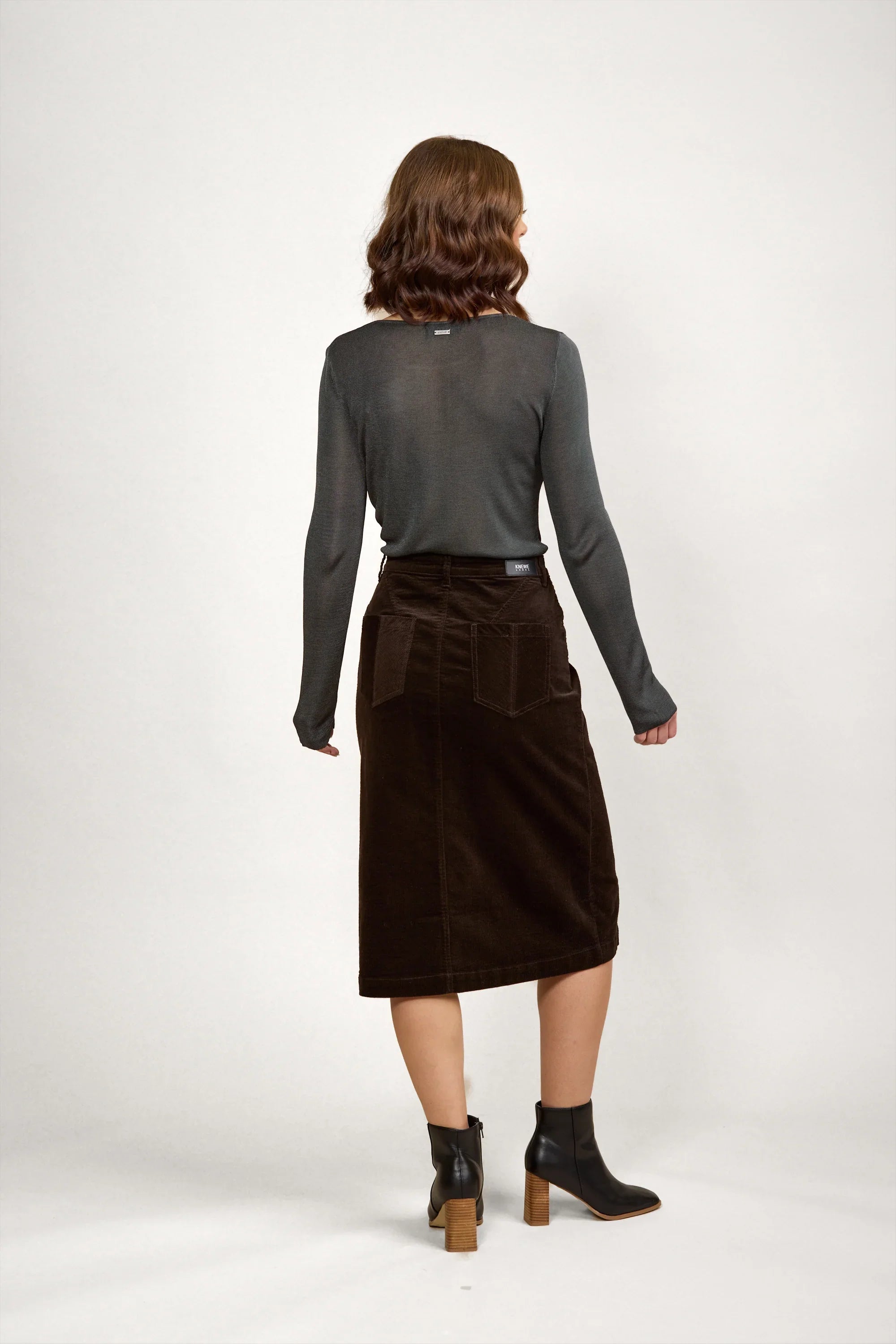 Woman wearing a dark corduroy midi skirt and long sleeve top with ankle boots, back view