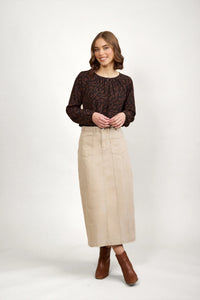 Woman in patterned brown blouse, beige midi denim skirt, and brown ankle boots against white background