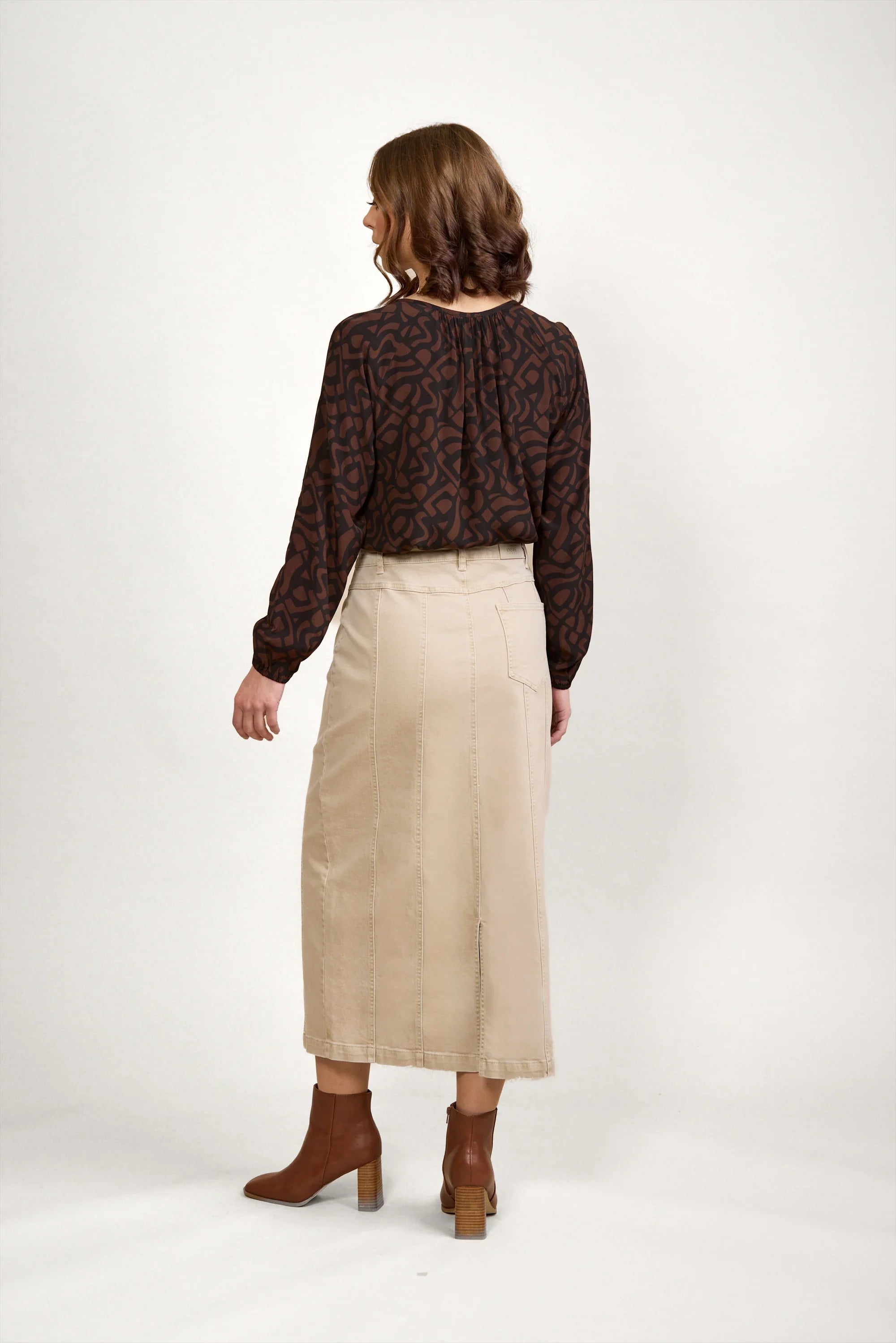 Woman wearing an oat denim midi skirt, brown abstract blouse, and ankle boots, back view.
