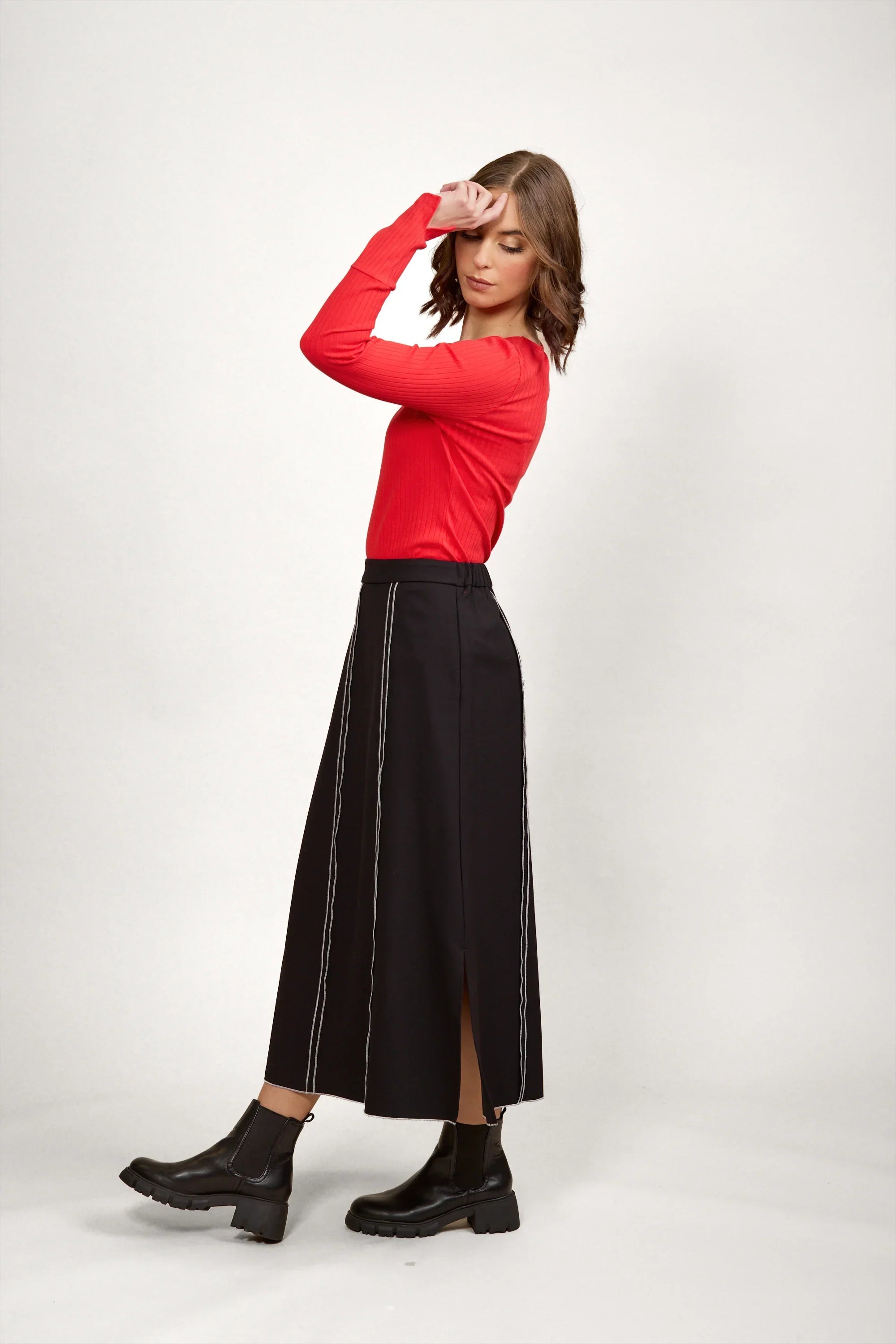 Woman in black midi skirt with white stitching, red long sleeve top, and black ankle boots