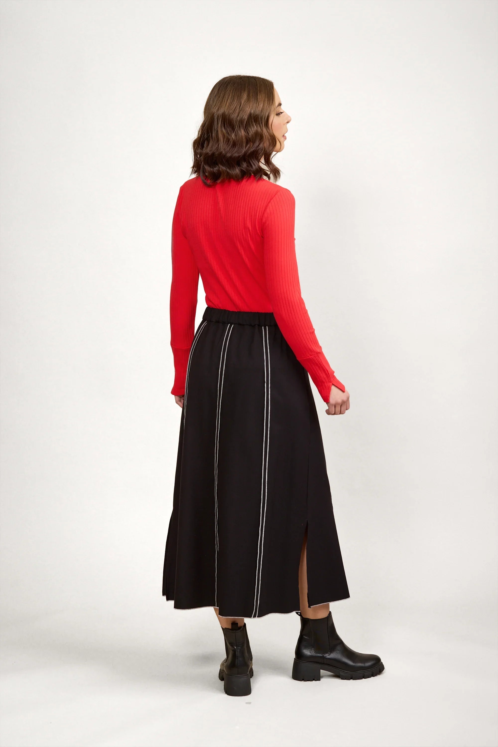 Woman wearing a black midi skirt with white vertical stitching, red ribbed top, black boots