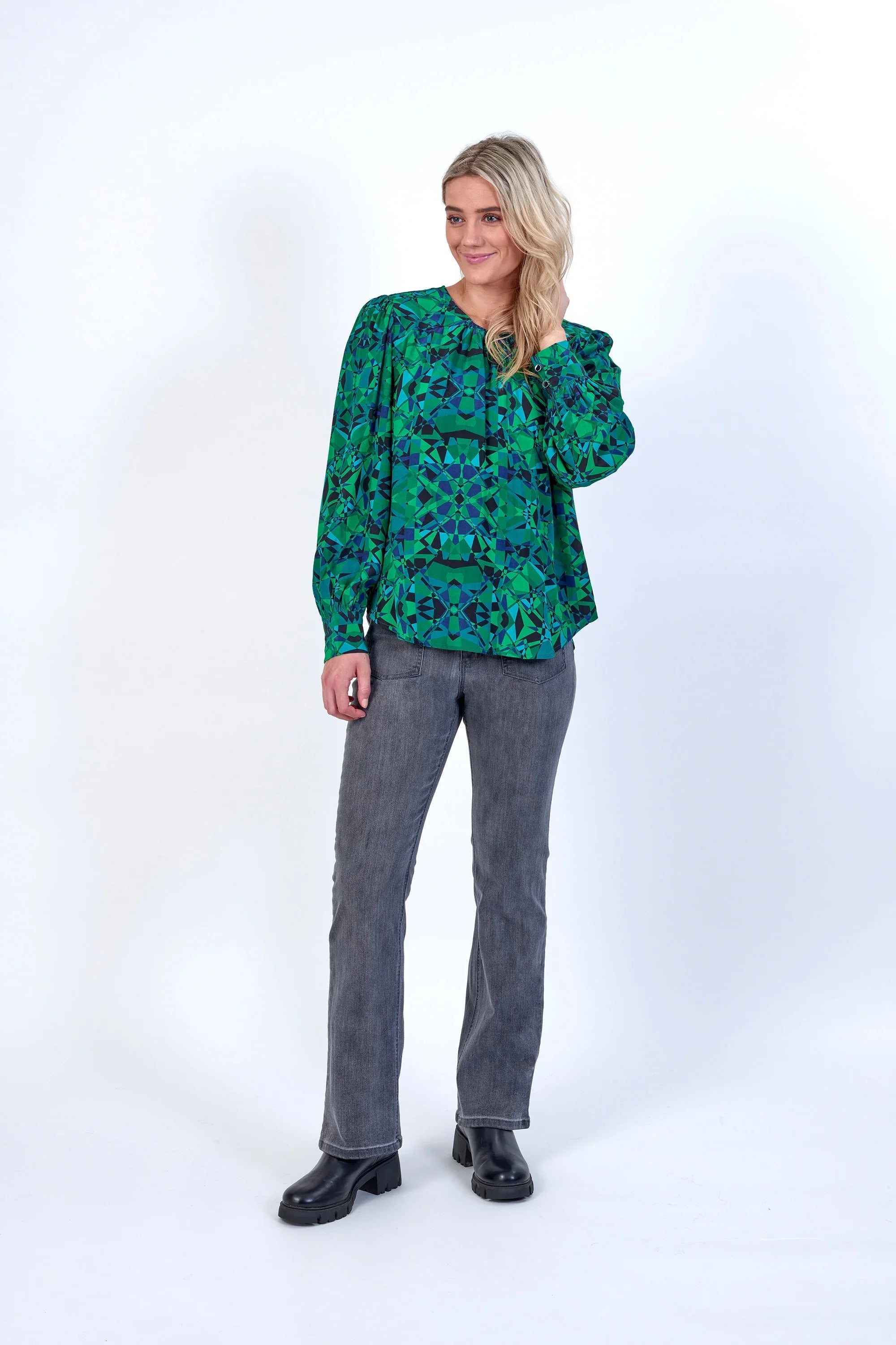 Woman in green geometric print long sleeve top, grey jeans, and black boots standing against a white background
