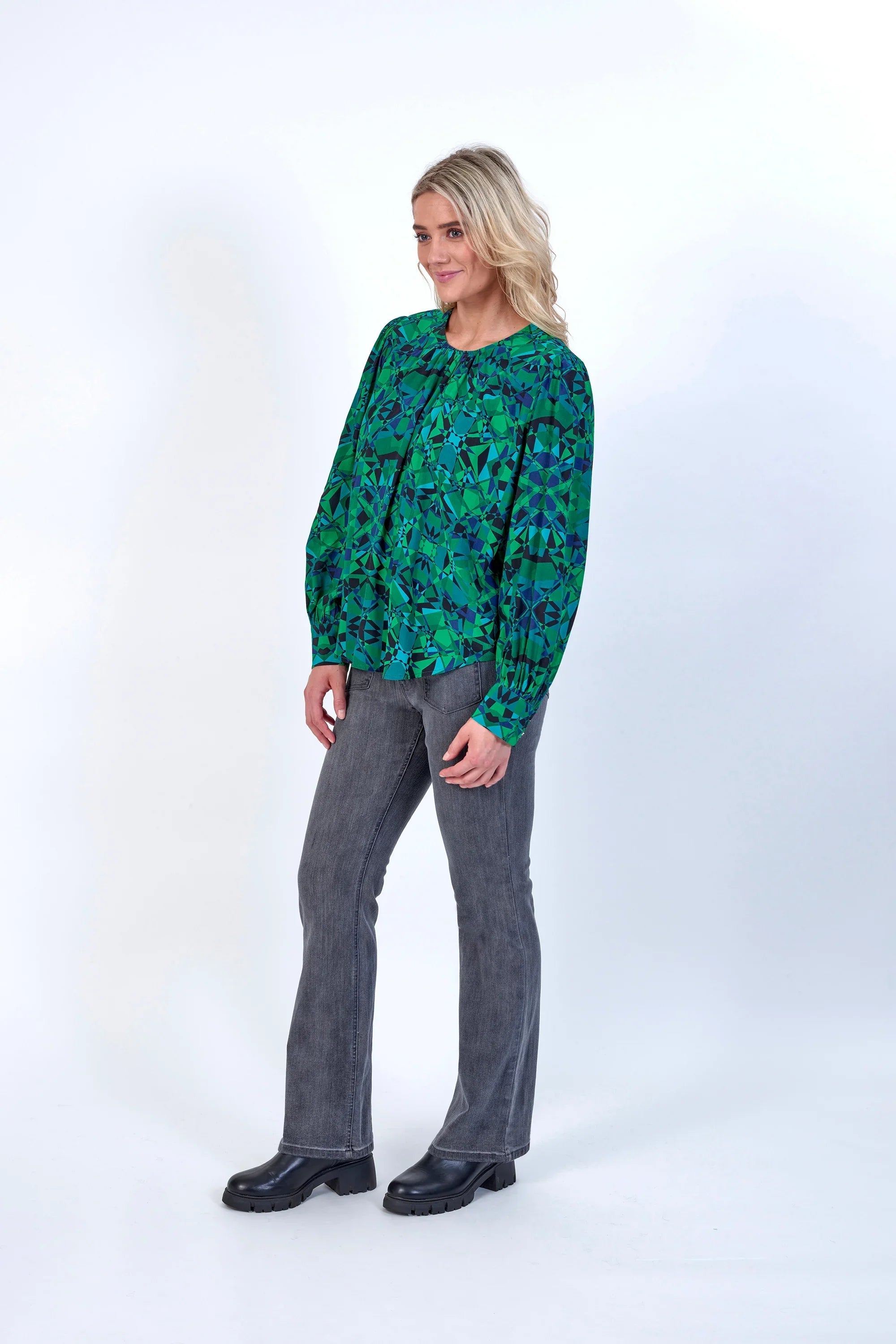 Woman wearing green patterned long sleeve blouse with grey jeans and black boots, studio background