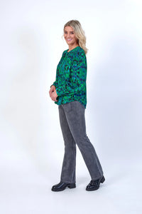 Woman in green geometric print blouse, grey jeans, and black boots standing against white background