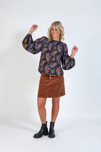 Woman in long-sleeve floral blouse, brown corduroy skirt, and black ankle boots standing against white background