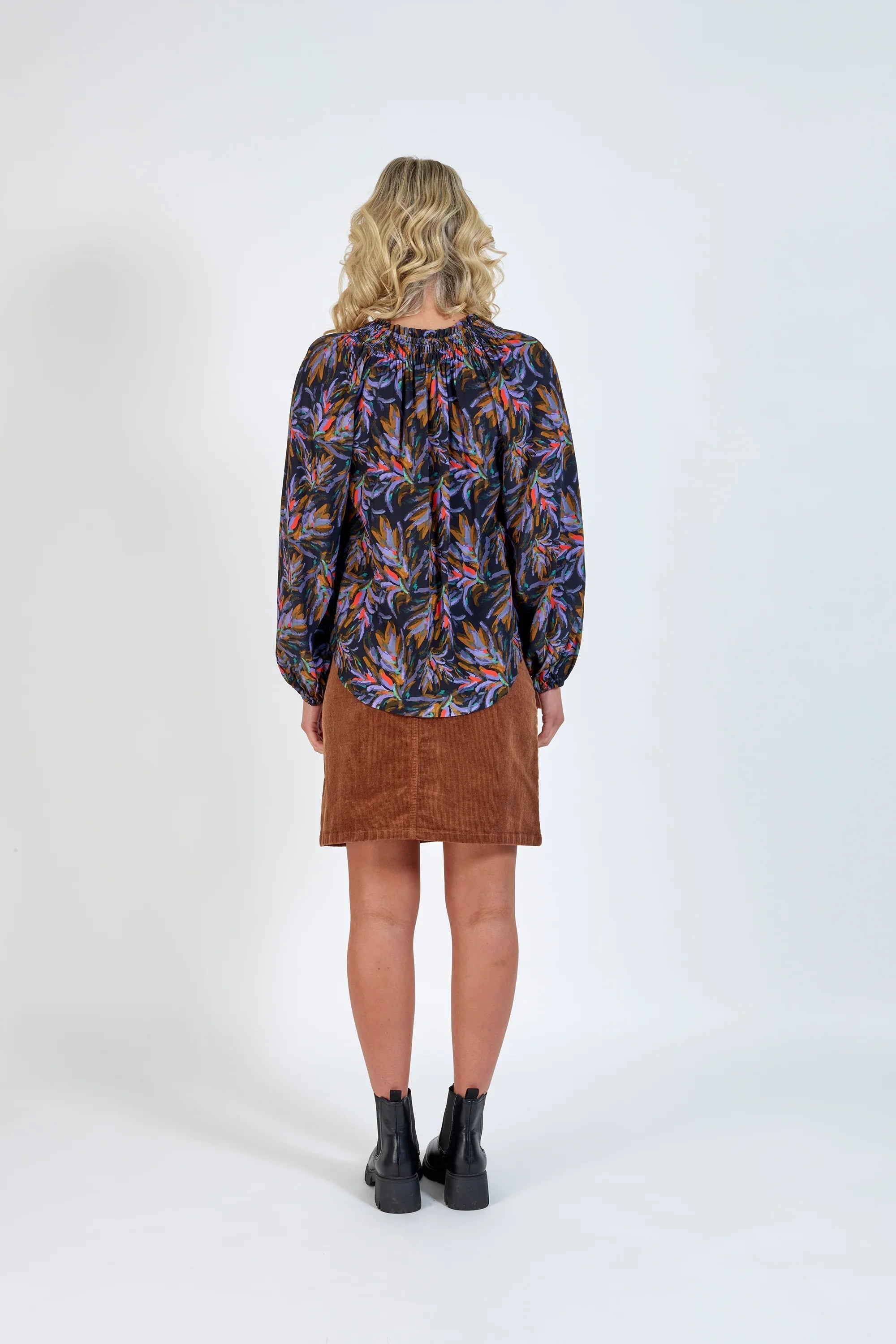 Woman in a colorful floral print blouse, brown corduroy skirt, and black ankle boots, back view