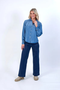 Woman wearing a relaxed blue denim button-up shirt and wide-leg jeans on white background