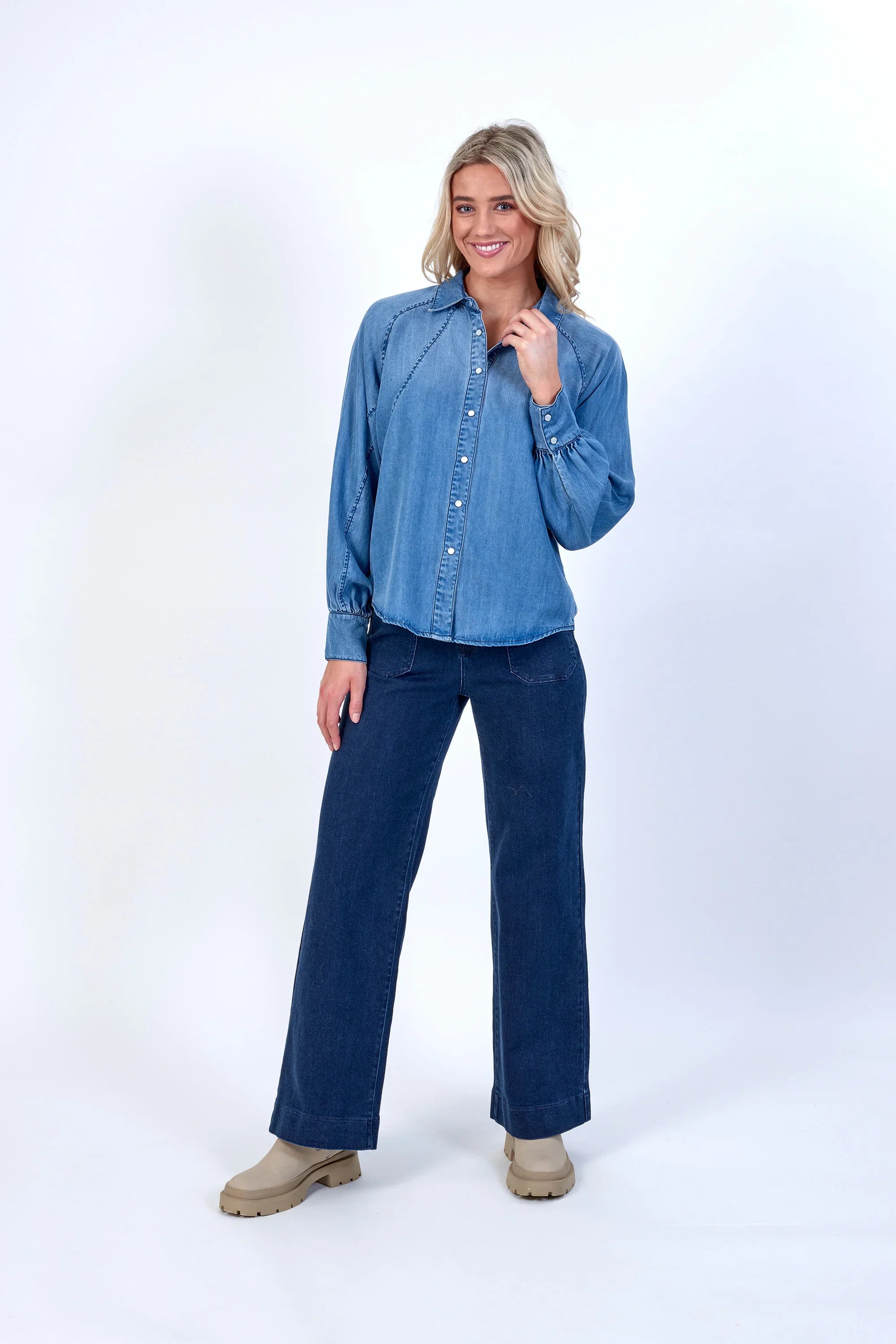 Woman wearing a relaxed blue denim button-up shirt and wide-leg jeans on white background
