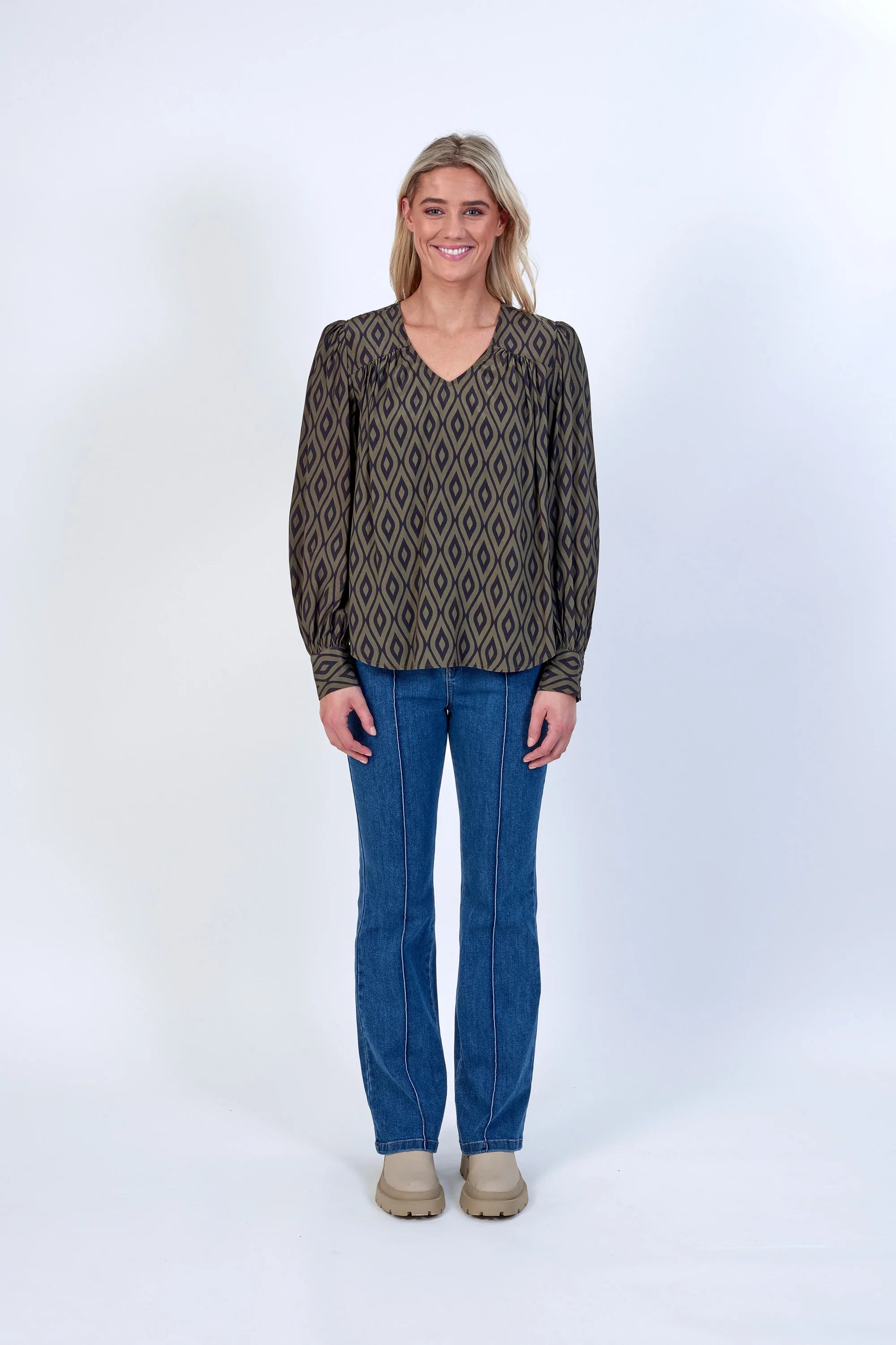Woman in geometric patterned long sleeve blouse, blue jeans, and beige boots, studio background