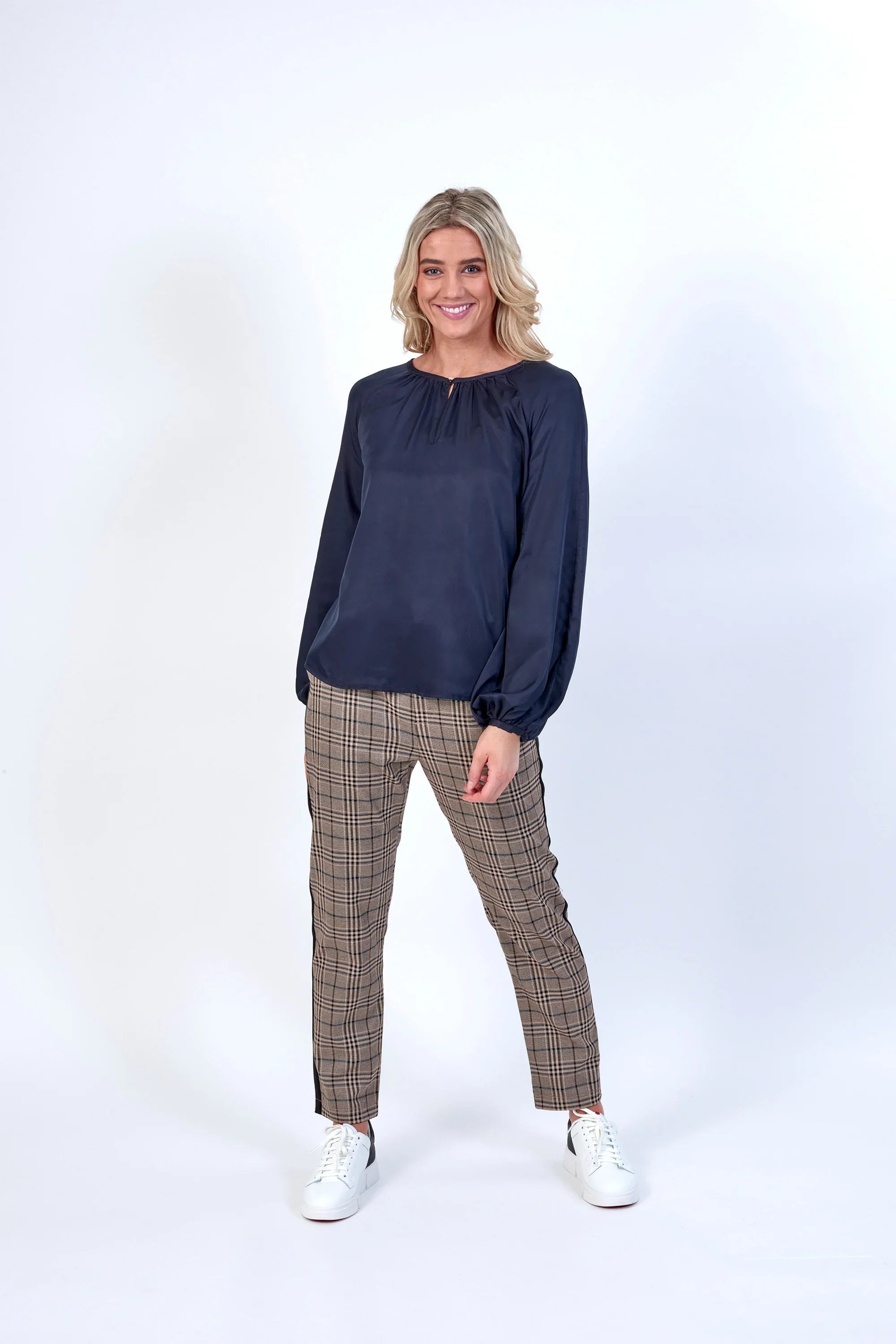 Woman wearing a navy long sleeve blouse and plaid pants with white sneakers, studio background
