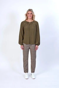 Model wearing a khaki long sleeve blouse with plaid trousers and white sneakers against a white background.