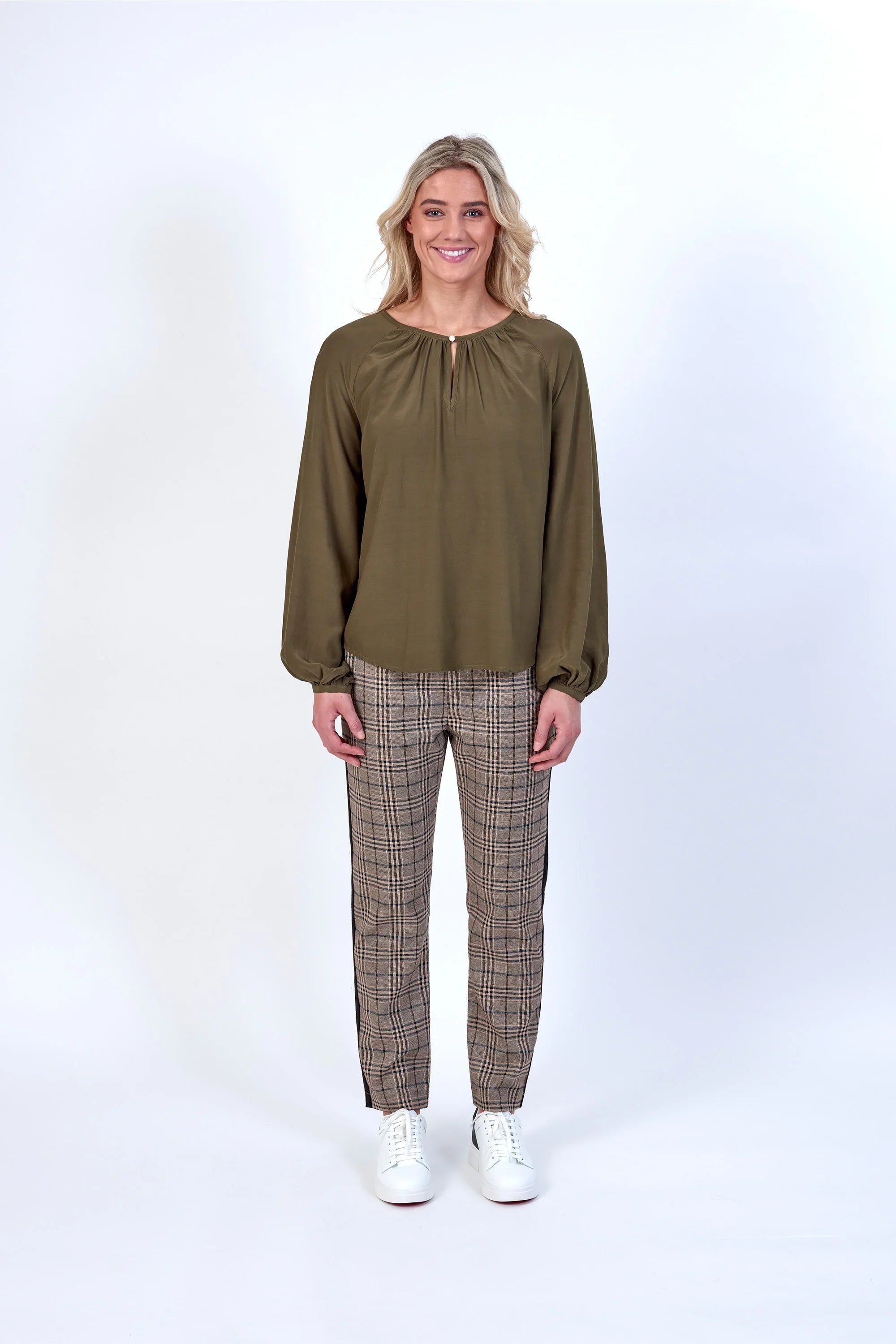 Model wearing a khaki long sleeve blouse with plaid trousers and white sneakers against a white background.