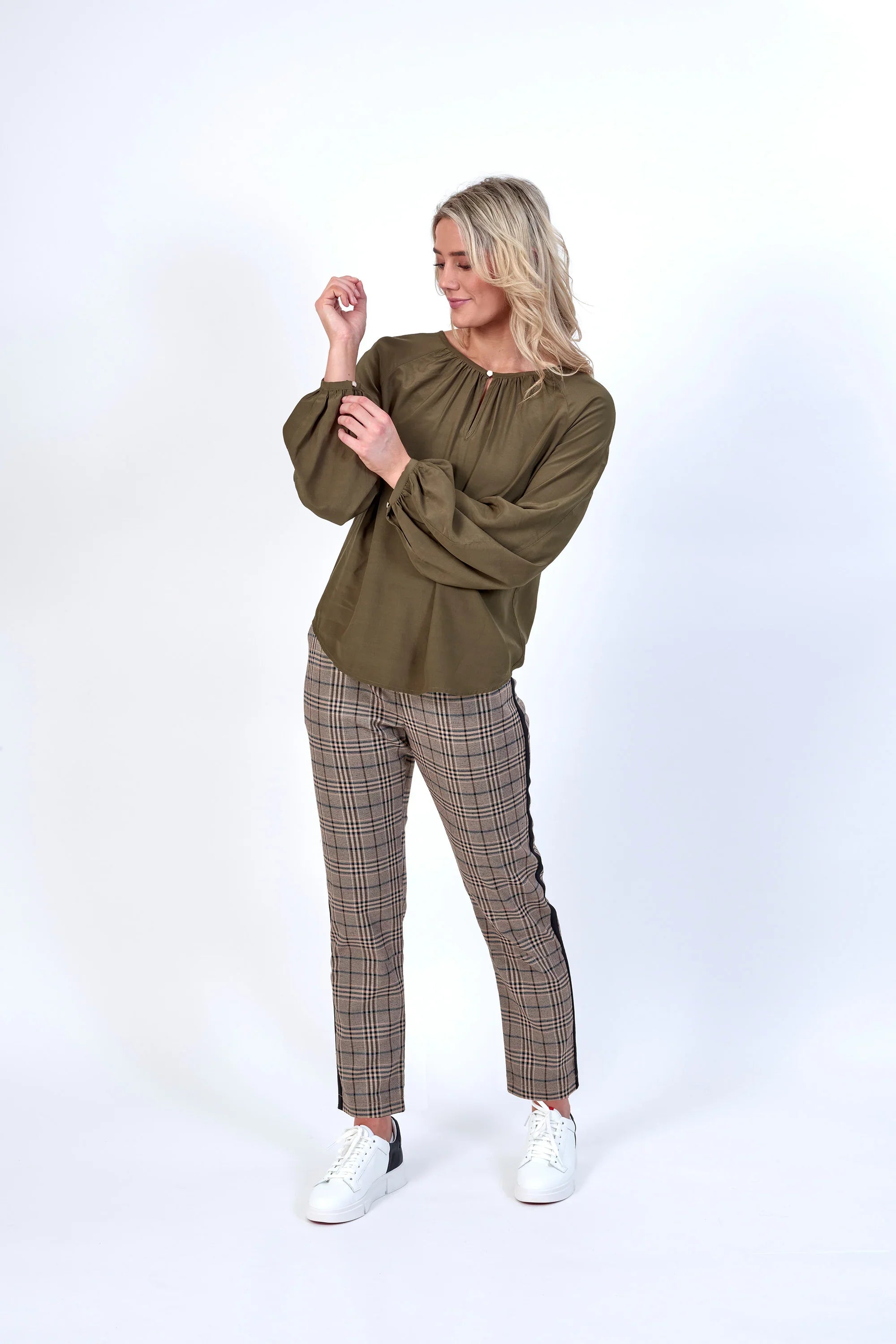 Woman wearing khaki long-sleeve blouse and plaid trousers with white sneakers, studio setting