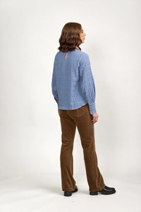 Woman in textured blue long sleeve top and brown pants, back view, studio setting