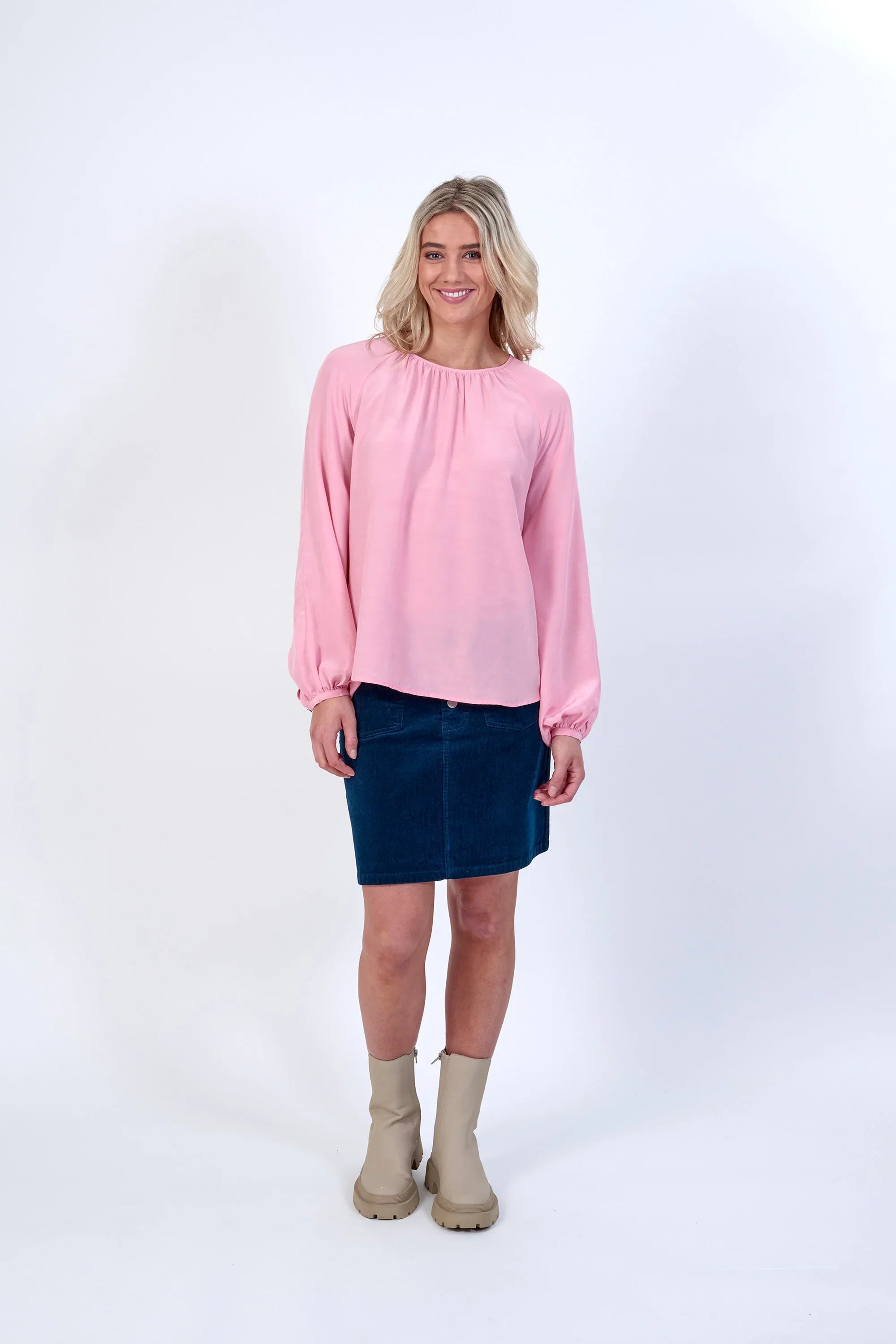 Woman wearing a light pink long sleeve top, blue skirt, and beige boots on white background